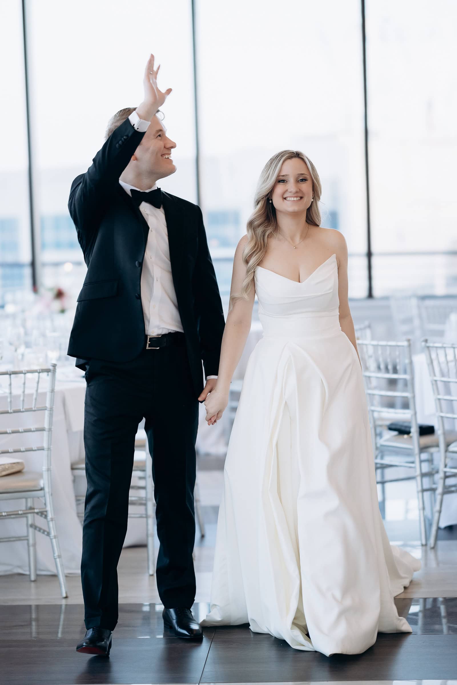 Bride in strapless white gown and groom in black tuxedo holding hands, walking indoors at Hudson House, Jersey City wedding venue.