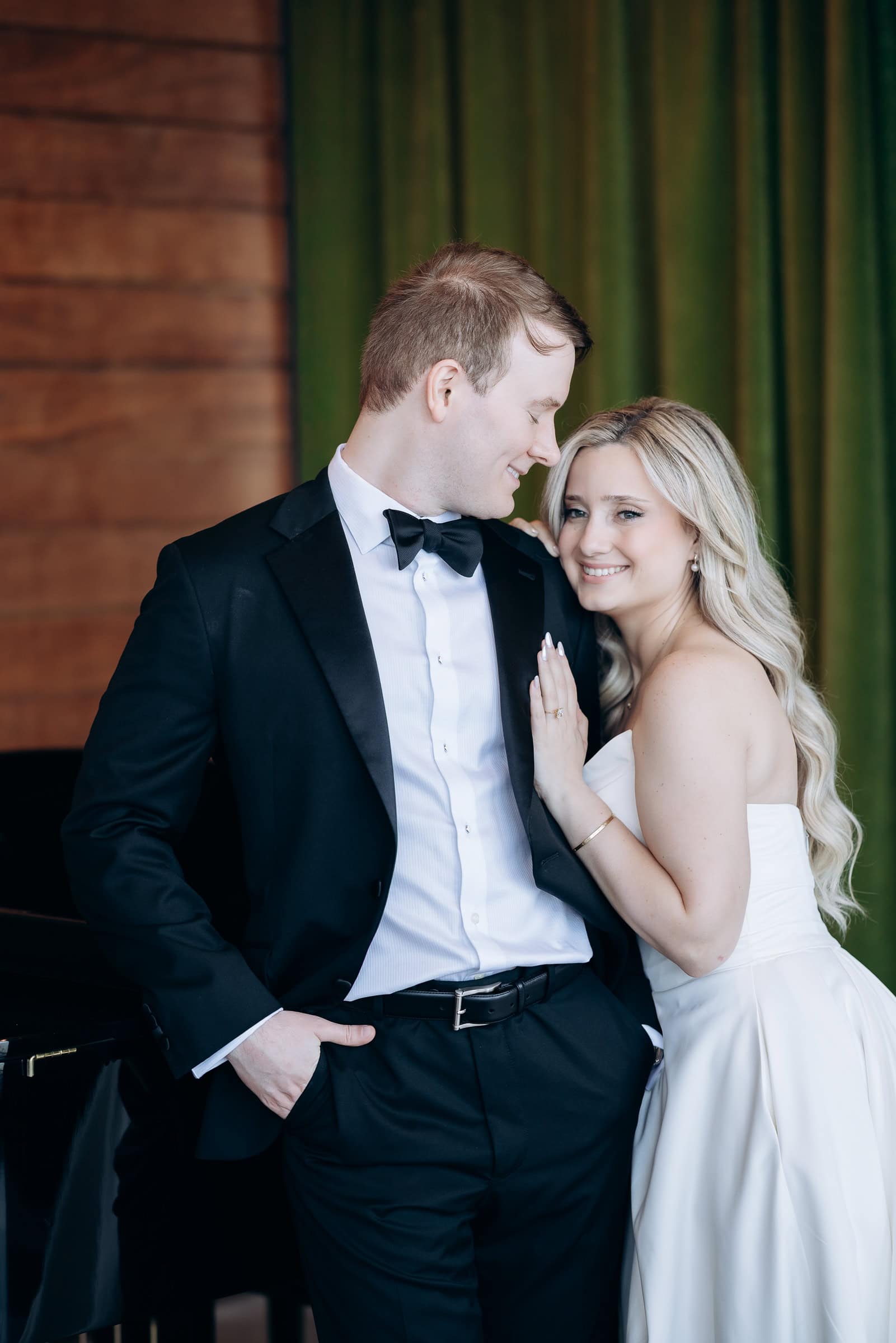 Groom in black tuxedo and bride in strapless white gown embracing, with green curtains and wooden wall background at Hudson House, Jersey City.