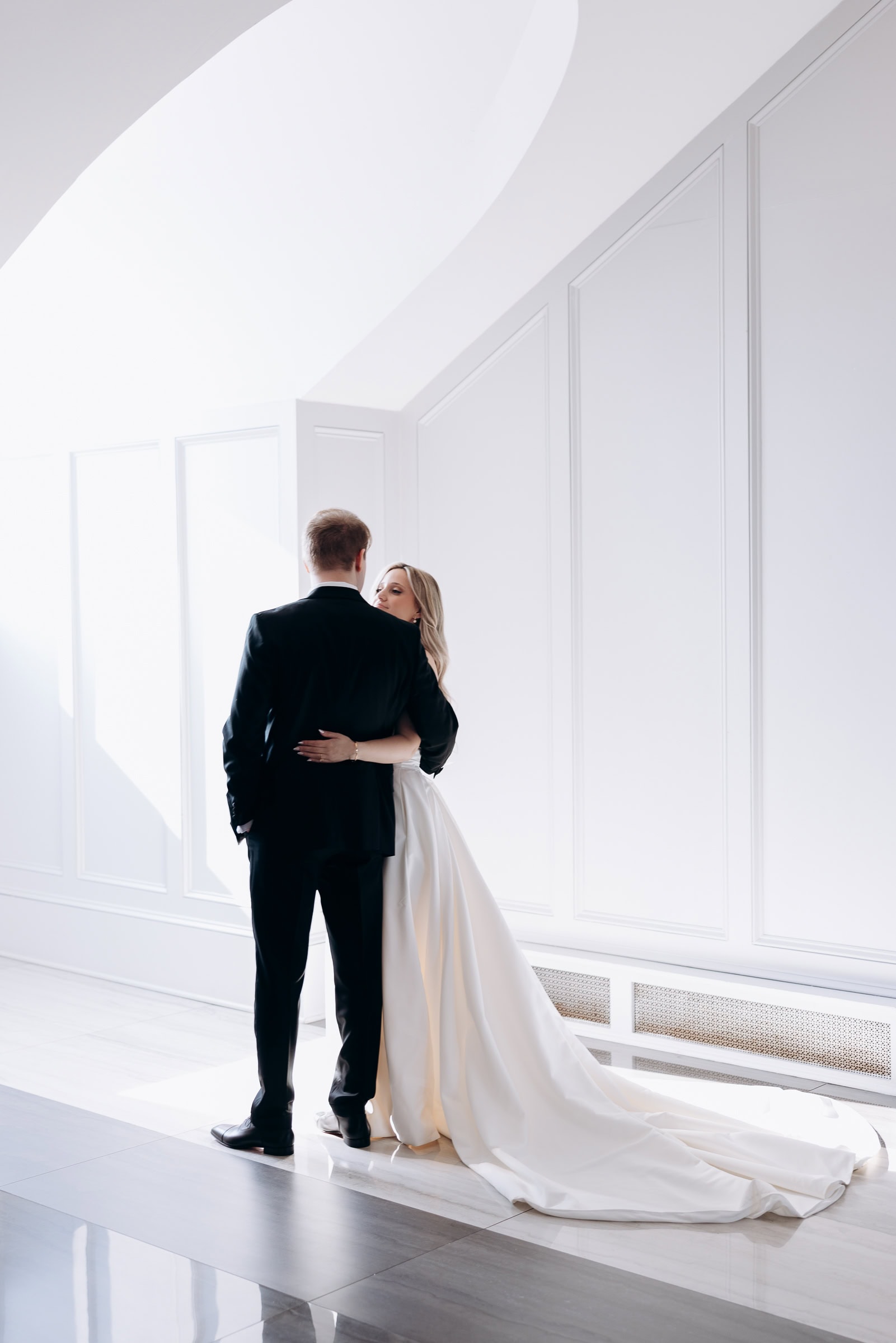 Bride and groom embracing near a large arched window in a bright, minimalist white interior at Hudson House, Jersey City, NJ.