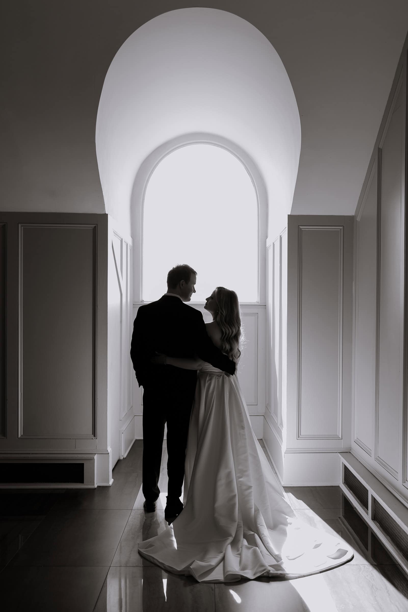 Bride and groom embracing in a bright arched corridor with white paneled walls and reflective marble floor at Hudson House, Jersey City, NJ.