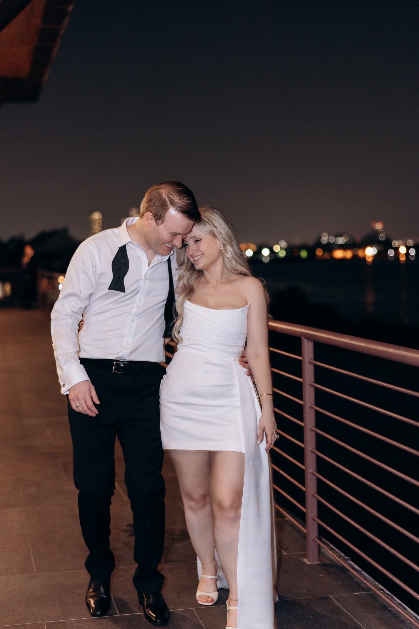 Couple in formal attire embracing on a balcony at night with blurred city lights in the background at Hudson House, Jersey City.
