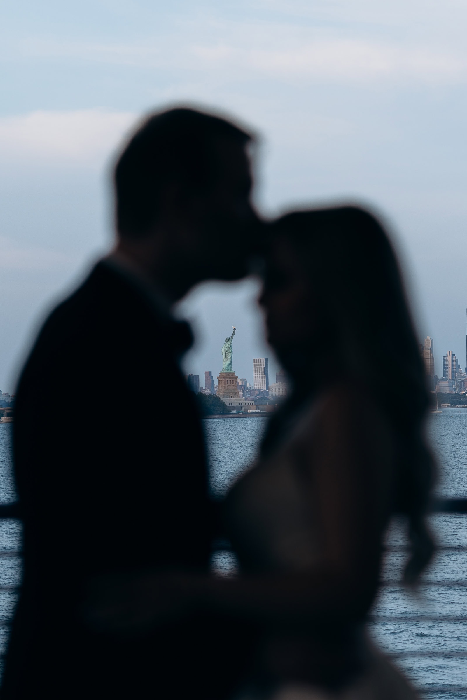Silhouetted couple in an embrace with the Statue of Liberty and Manhattan skyline in soft evening light behind them.