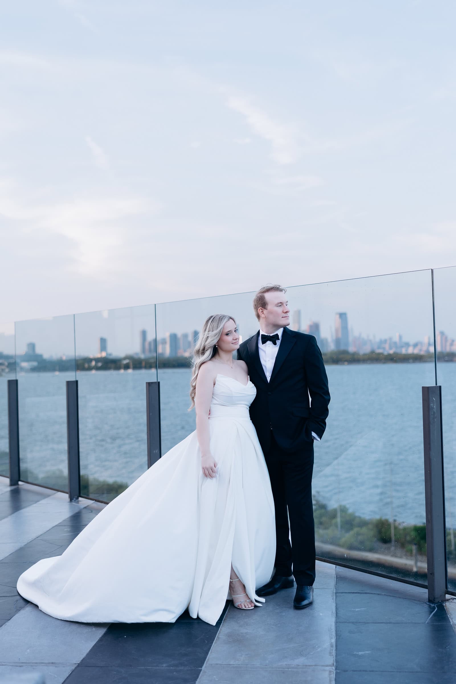 Bride in white strapless gown and groom in black tuxedo standing by glass railing overlooking Hudson River and Manhattan skyline at sunset.