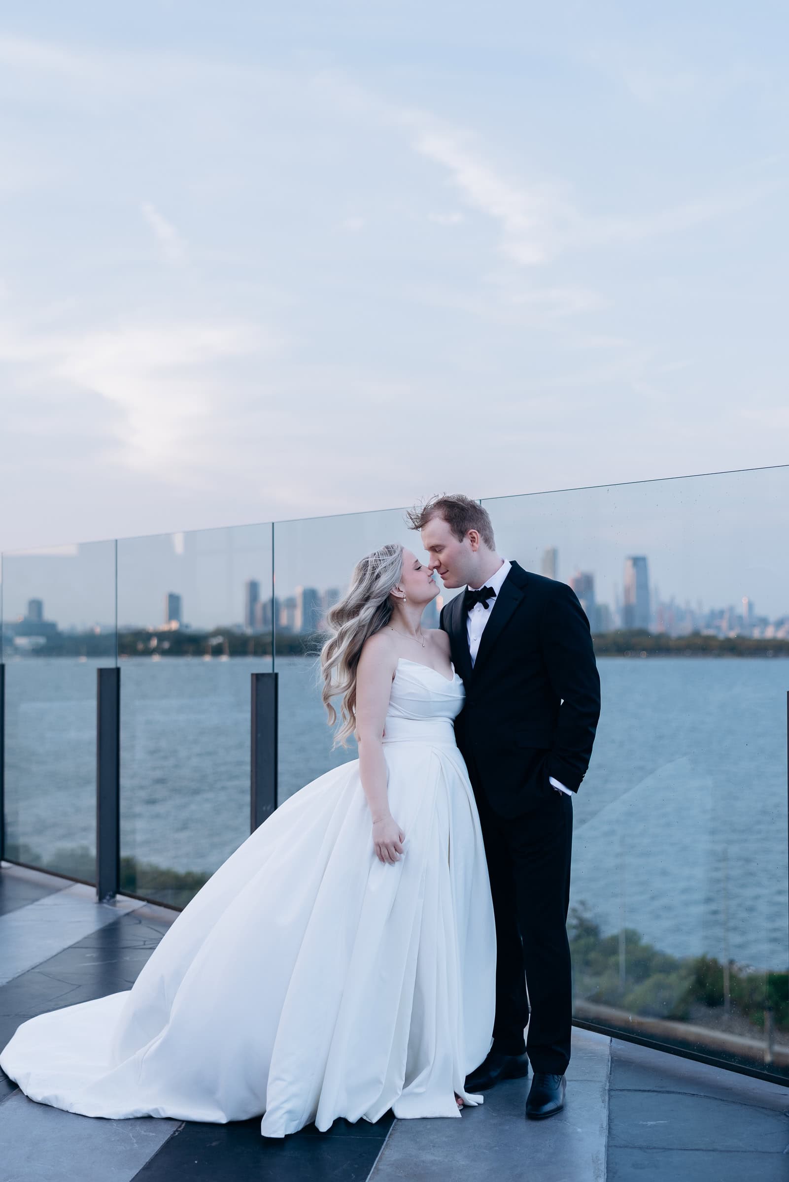 Bride in a voluminous white gown and groom in black tuxedo share an intimate moment on a rooftop terrace with Manhattan skyline and water in the background.