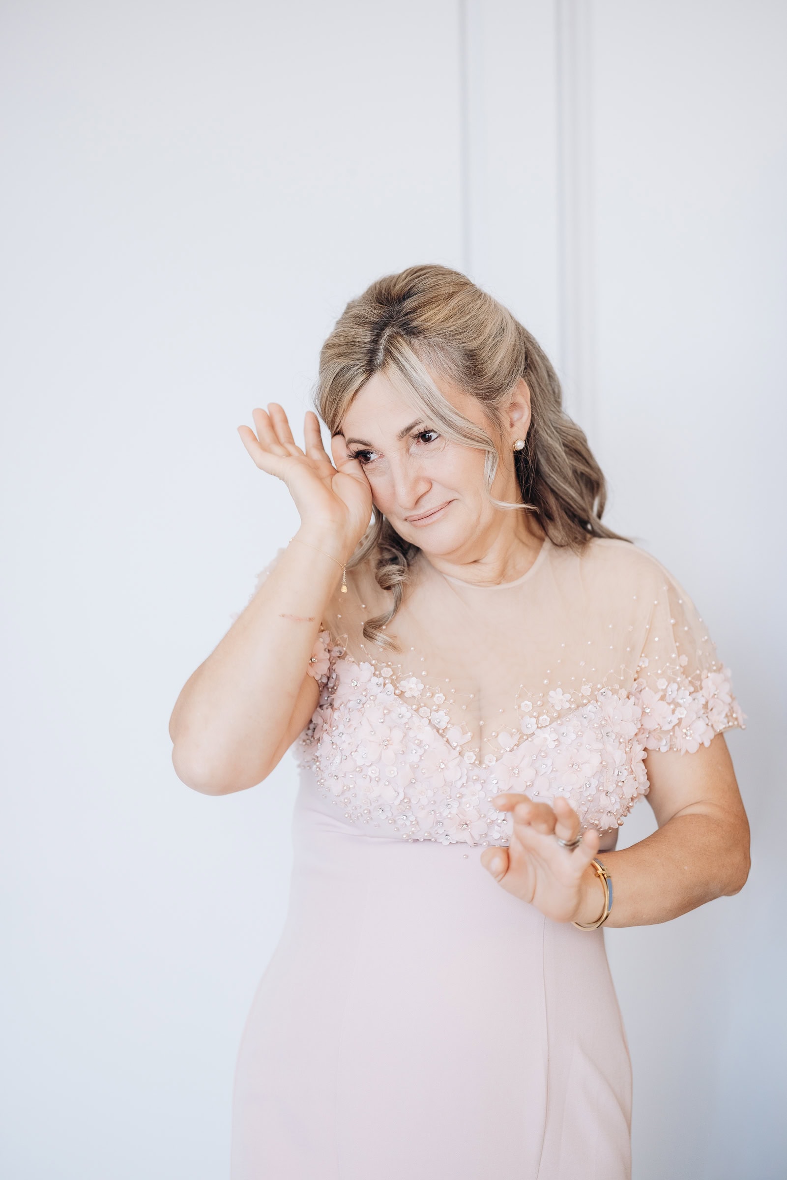 Woman in blush dress with floral embellishments wiping a tear from her eye against a neutral background.
