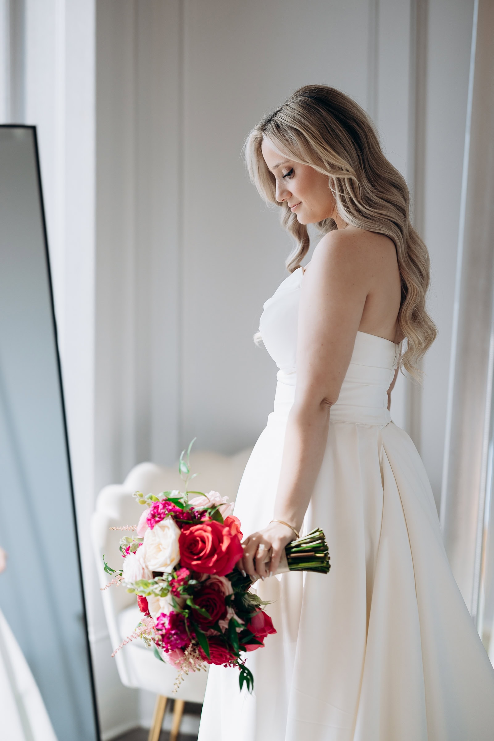 Bride in strapless ivory wedding gown holding vibrant bouquet of pink and cream roses, standing near window with soft natural light at Hudson House, Jersey City.