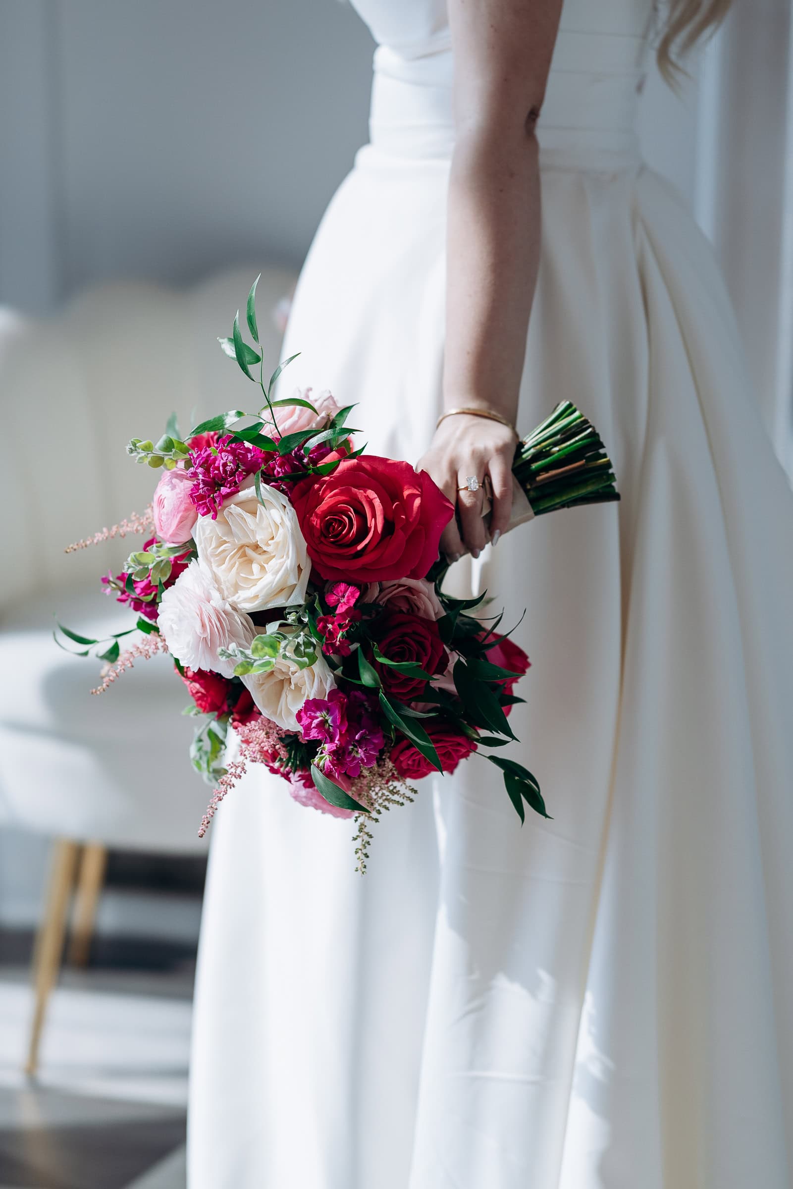 Bride in white strapless gown holding bouquet of red, pink, and cream roses with greenery, standing indoors near a mirror at Hudson House, Jersey City.