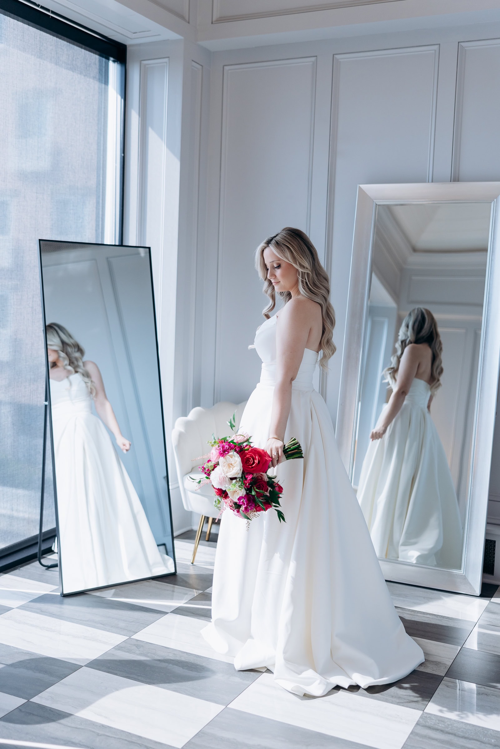 Bride in ivory strapless gown holding bouquet, reflected in two full-length mirrors in a sunlit room with checkered floor at Hudson House, Jersey City.