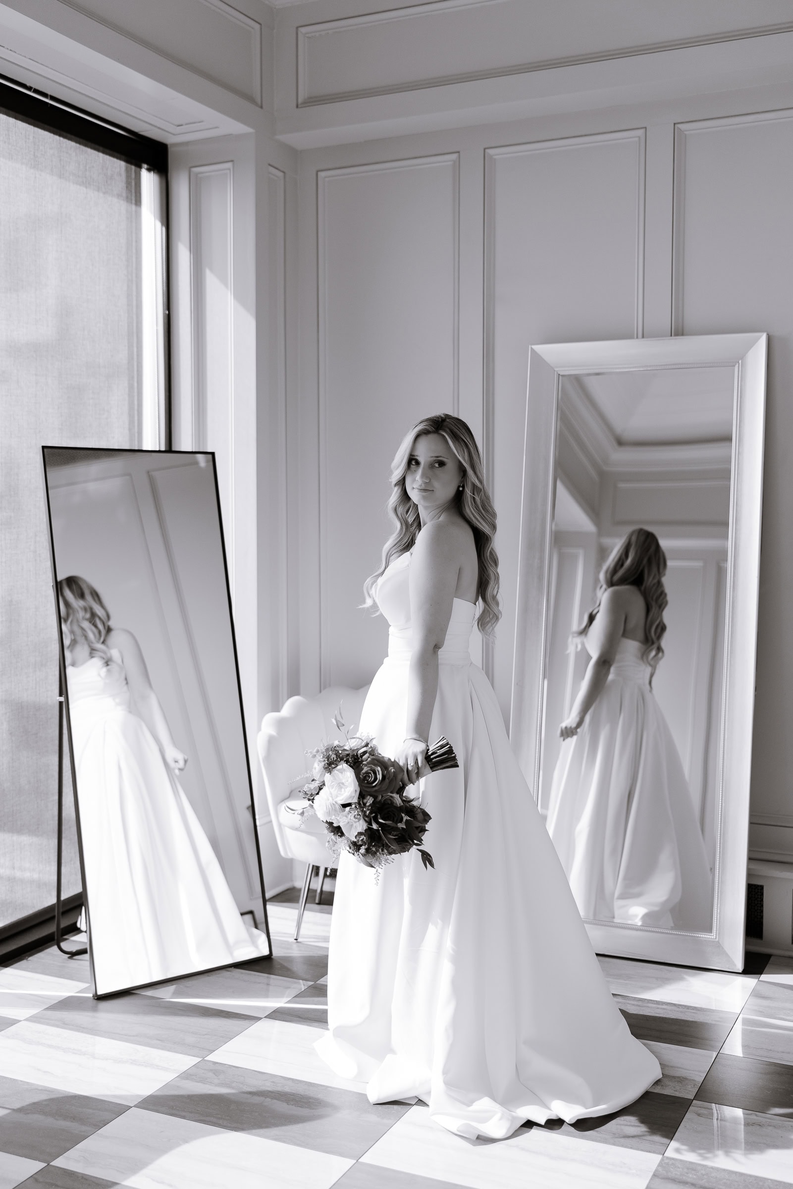 Bride in ivory strapless wedding gown holding pink and red bouquet, standing near two mirrors in a softly lit room at Hudson House, Jersey City.