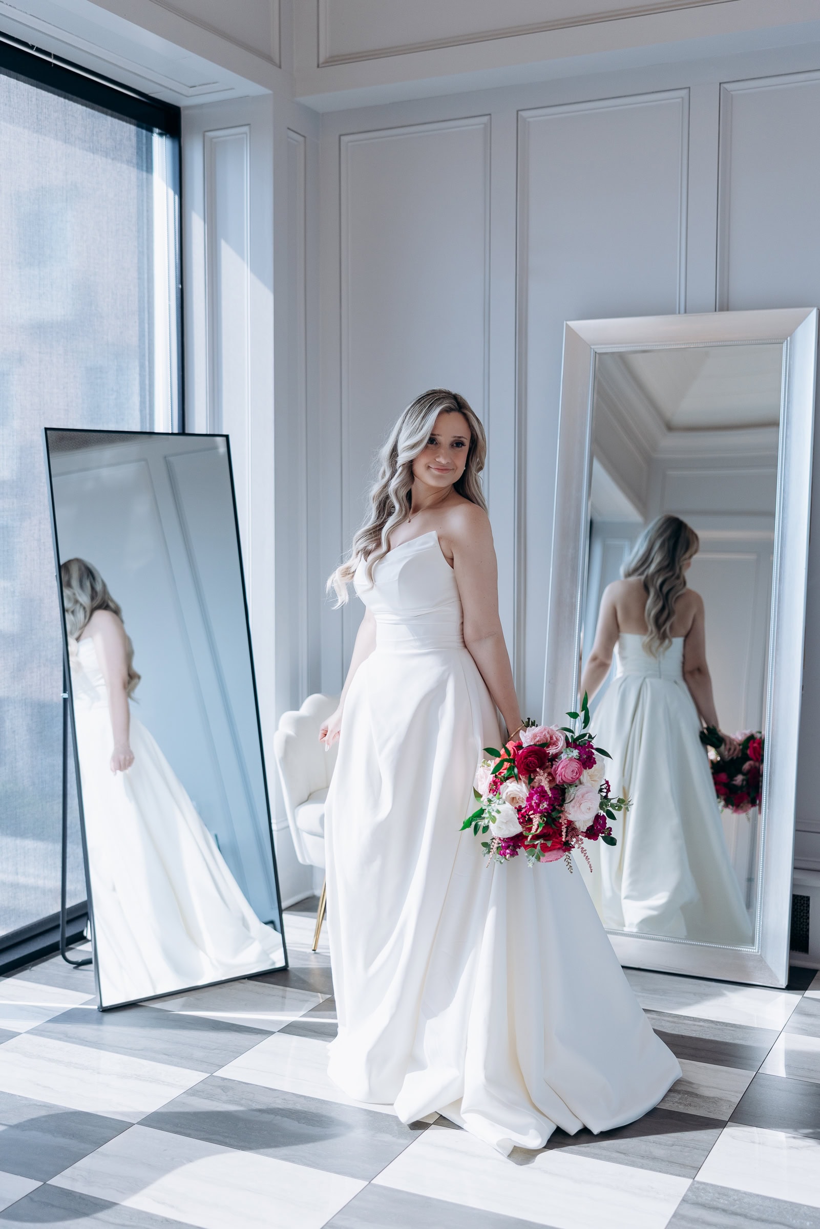 Bride in satin wedding dress holding pink and red floral bouquet standing between two full-length mirrors in a sunlit room with checkered marble floor at Hudson House, Jersey City.