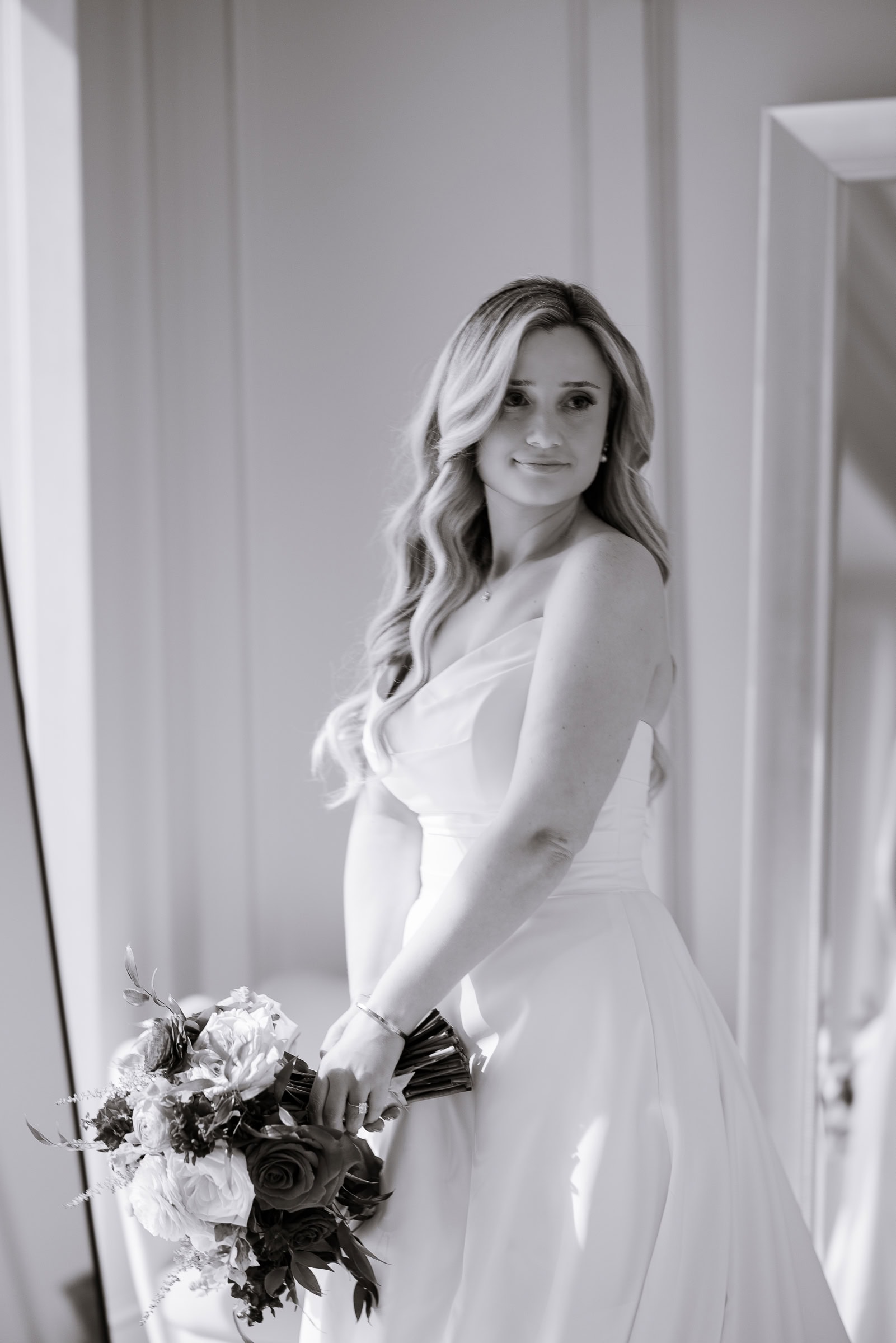 Bride in satin strapless gown holding vibrant bouquet of red and pink flowers, standing near window with soft light at Hudson House, Jersey City.