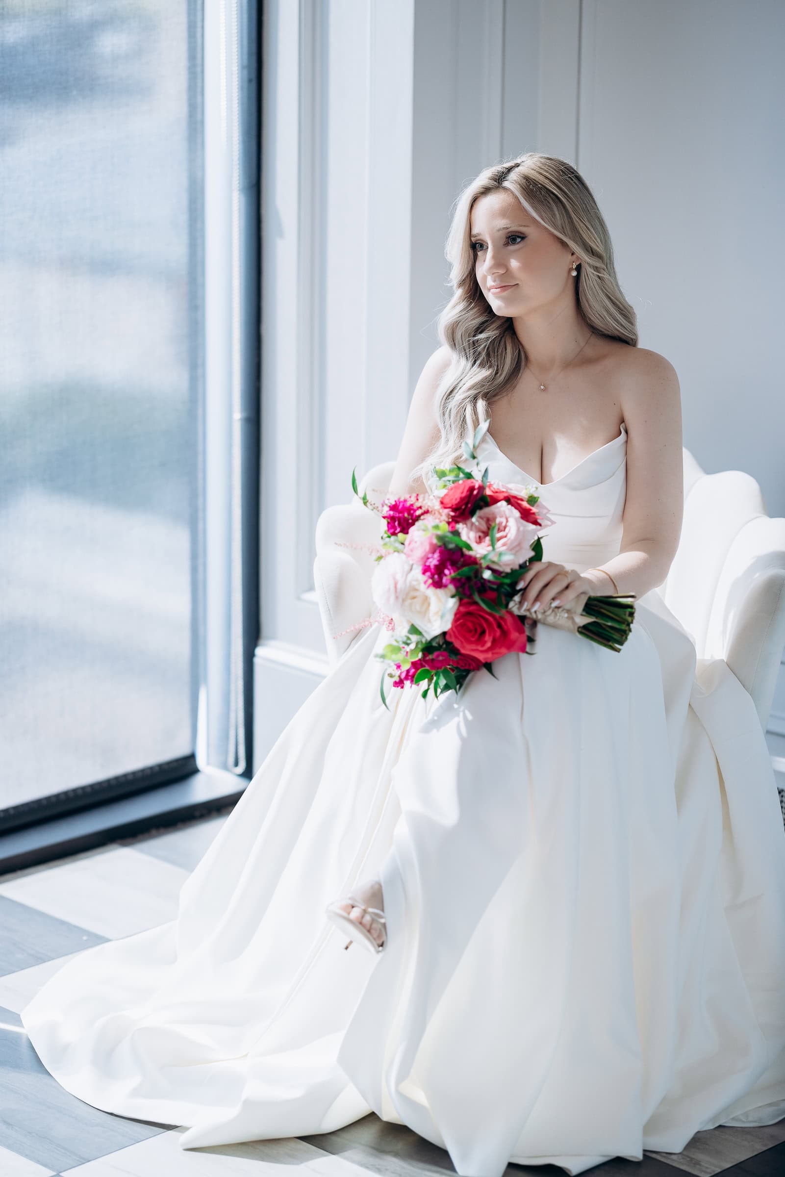 Bride in ivory sculptural wedding gown seated by window holding vibrant red, pink, and white floral bouquet at Hudson House, Jersey City.