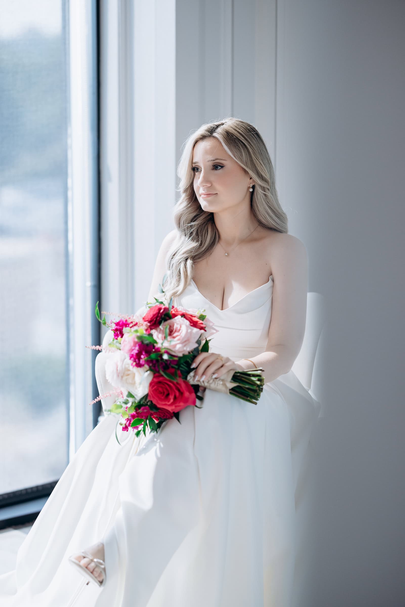 Bride in ivory strapless gown seated by window holding bouquet of red and pink flowers, soft natural light, Hudson House venue, Jersey City
