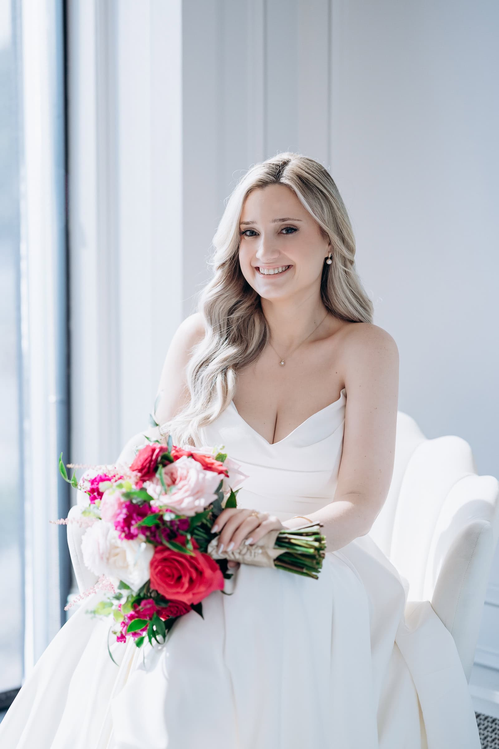 Bride in silk wedding dress seated in white chair holding vibrant pink and cream floral bouquet near window at Hudson House, Jersey City.
