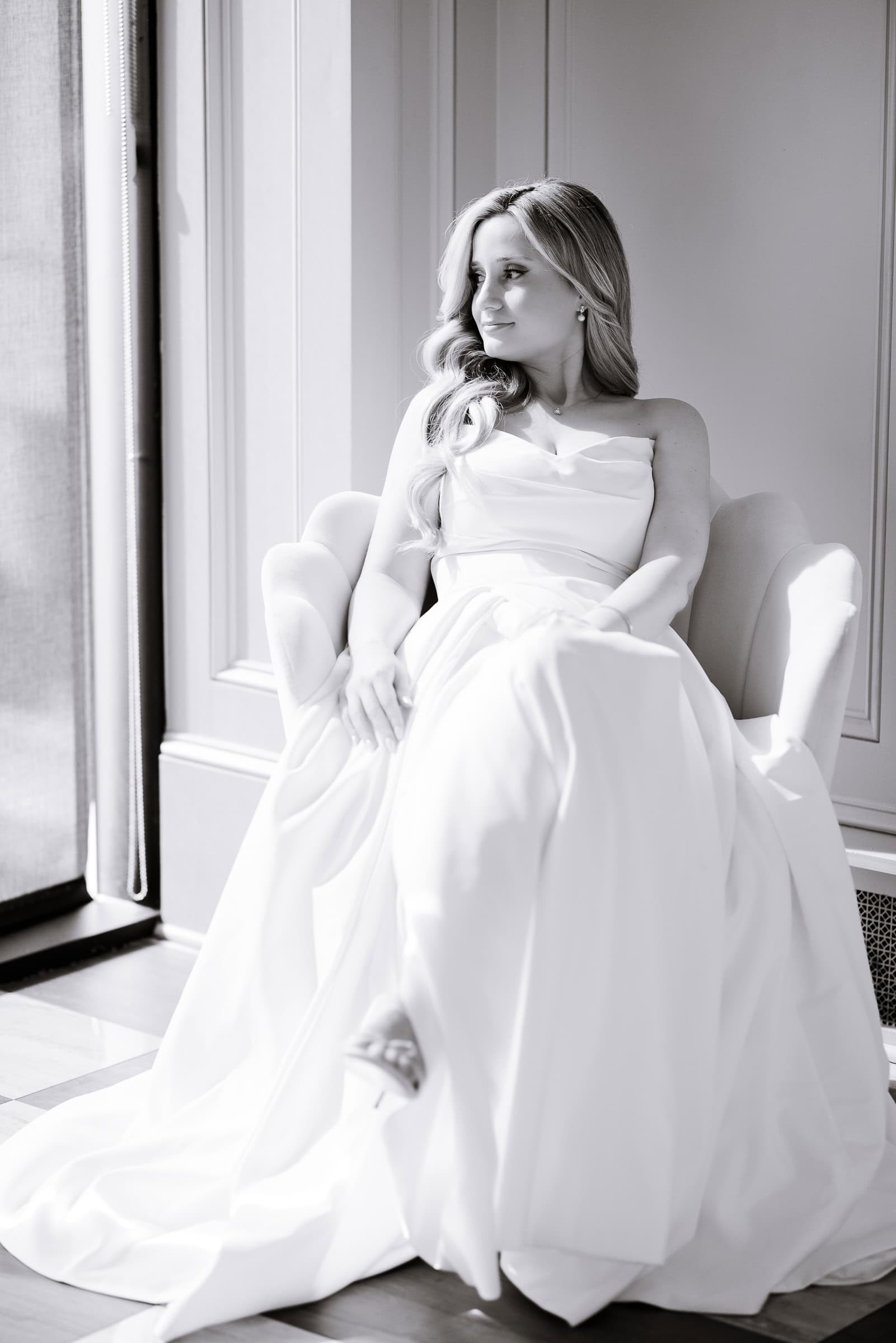 Bride in elegant silk wedding dress seated in a cream chair by a window, soft natural light illuminating her profile at Hudson House, Jersey City, NJ.