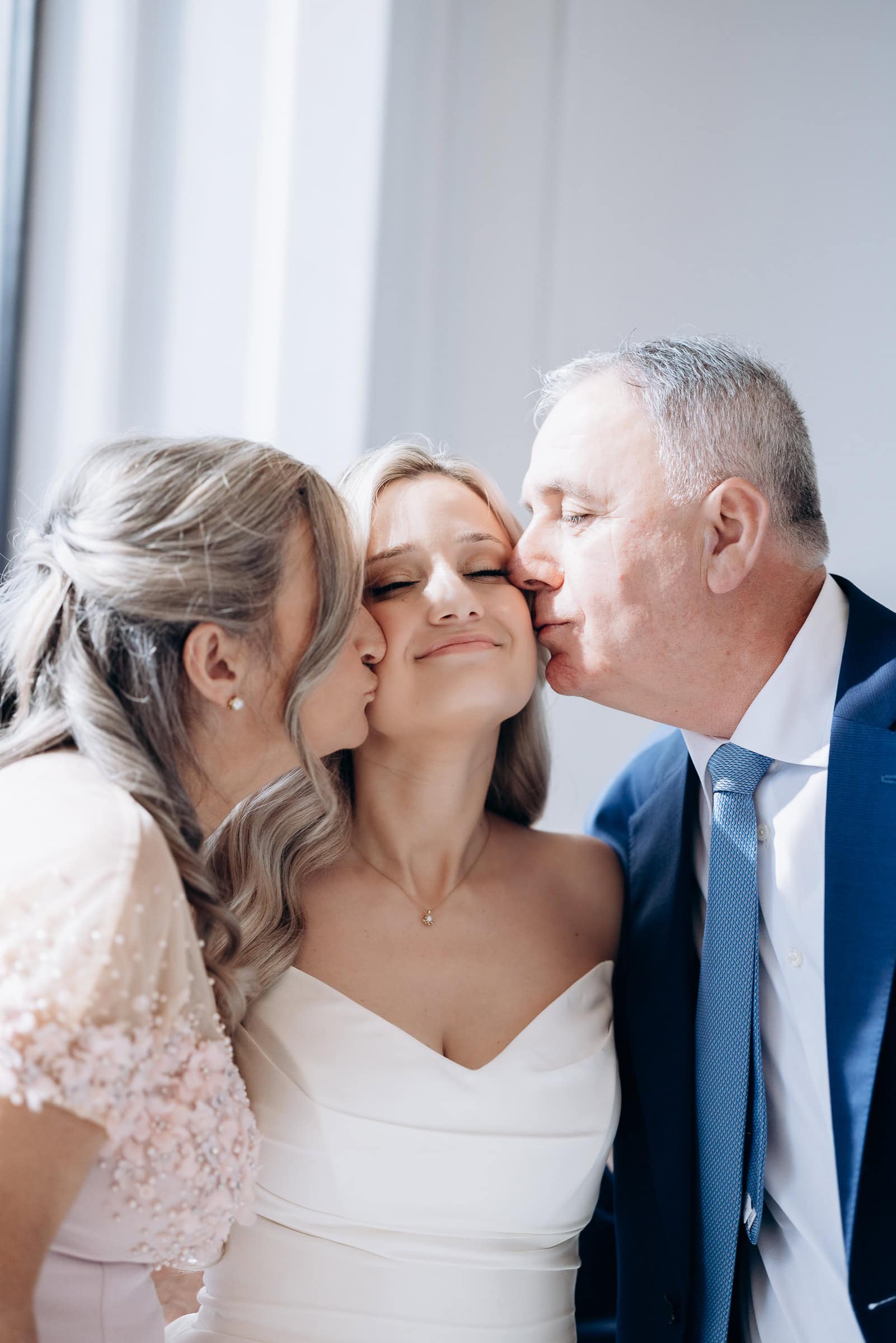 Bride in off-shoulder gown with eyes closed, kissed on both cheeks by parents in elegant attire, soft natural light indoors at Hudson House