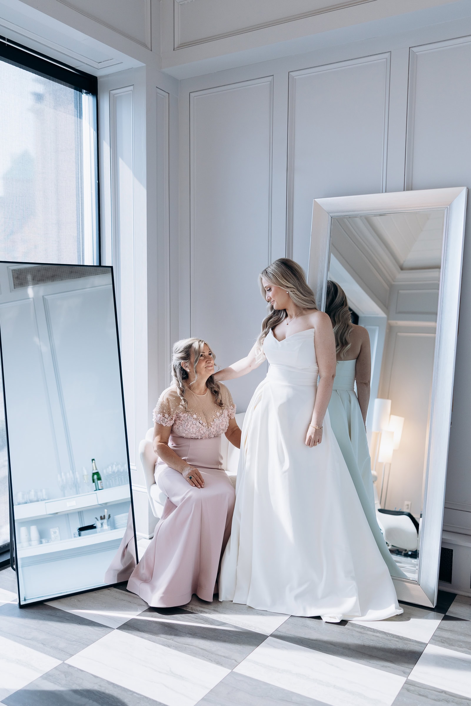 Bride in ivory gown standing beside seated woman in blush pink dress, reflected in tall mirror in elegant Hudson House room with checkered floor.
