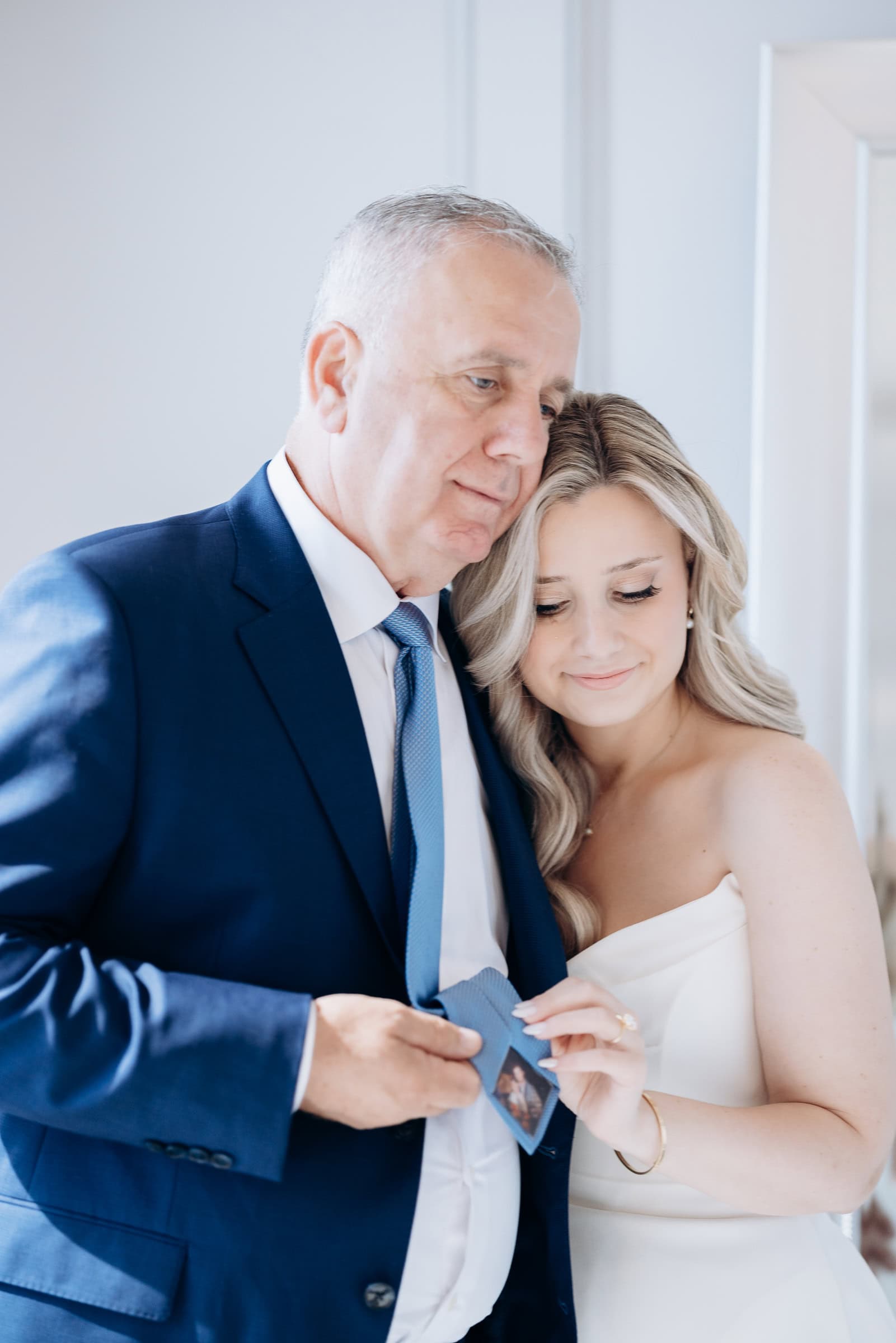 Bride and her father embracing closely, looking at a photo inside his blue tie in a softly lit room at Hudson House, Jersey City.