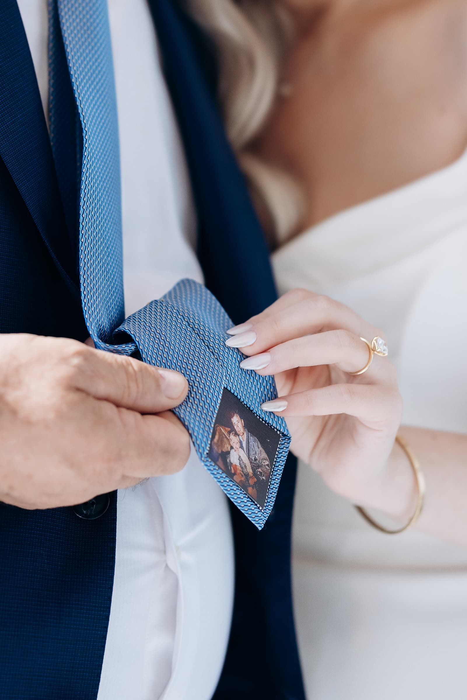 Close-up of a bride adjusting a blue tie with a photo patch on an older man in a navy suit, evoking a tender wedding moment.
