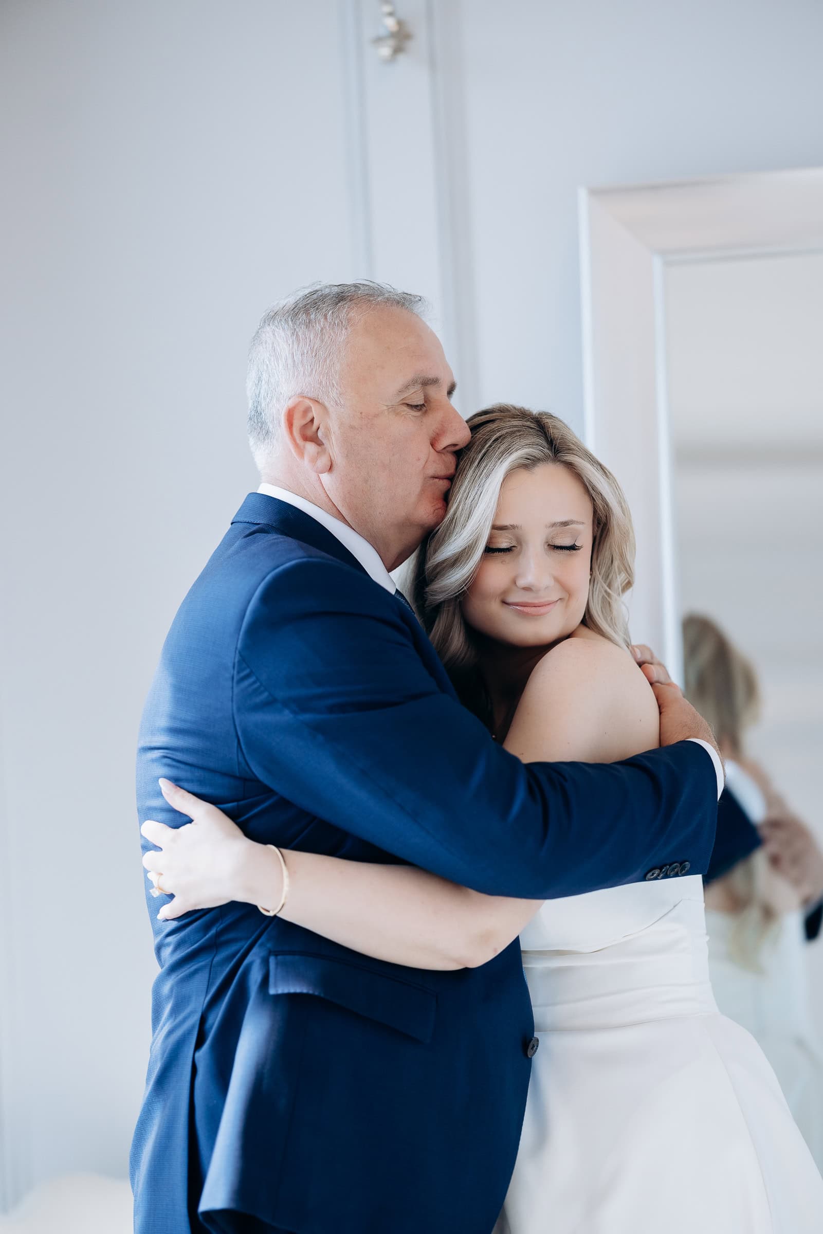 Father in navy suit tenderly embracing bride in white gown with eyes closed, soft natural light, Hudson House Jersey City
