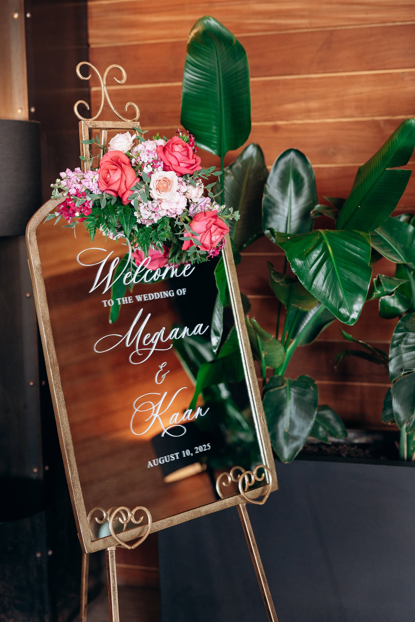Gold-framed mirror wedding welcome sign decorated with pink roses and greenery, set against wooden wall and large green leaves.