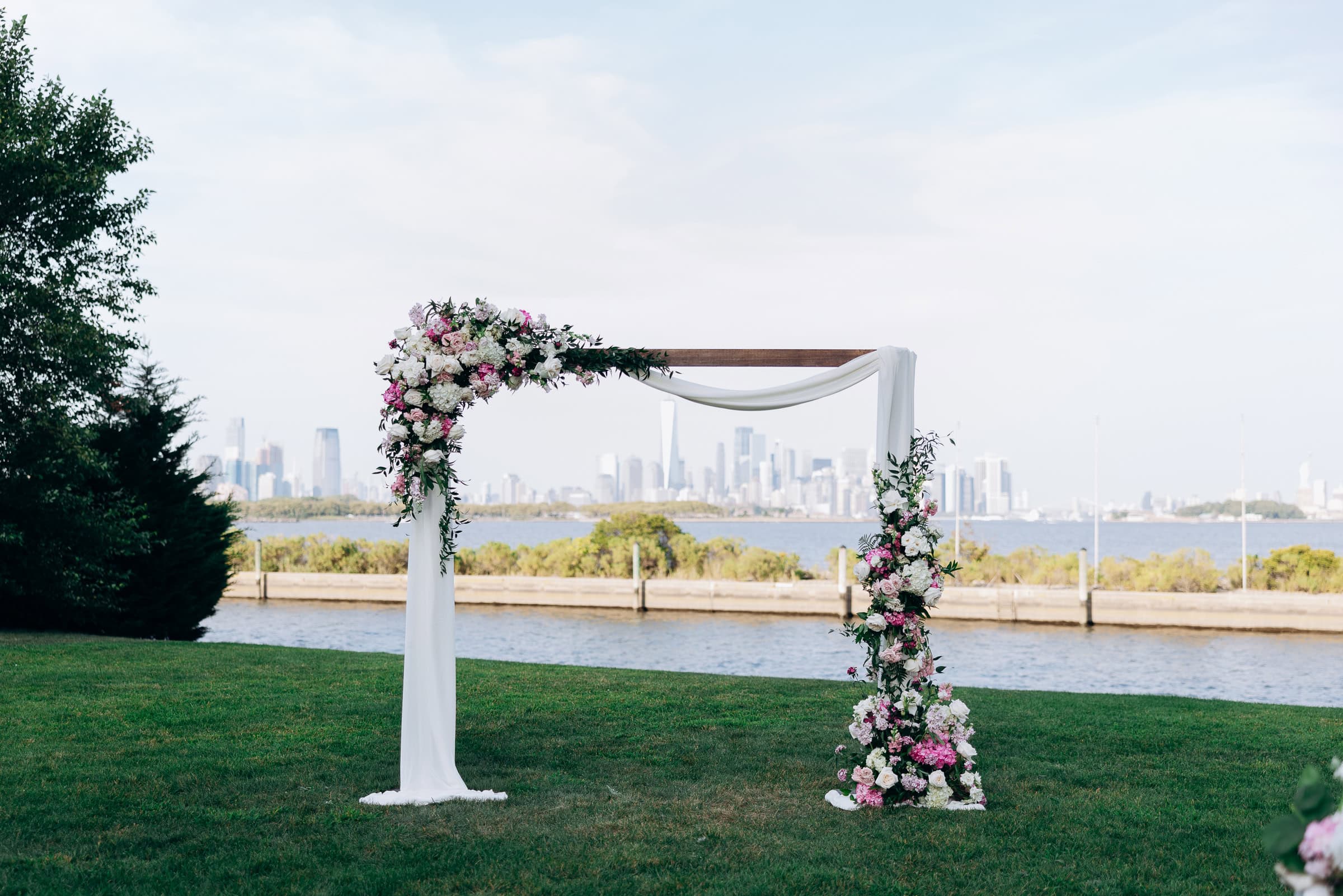 Wooden wedding arch decorated with white and pink flowers and white fabric, set on green lawn by waterfront with city skyline in the distance at Hudson House, Jersey City, NJ.