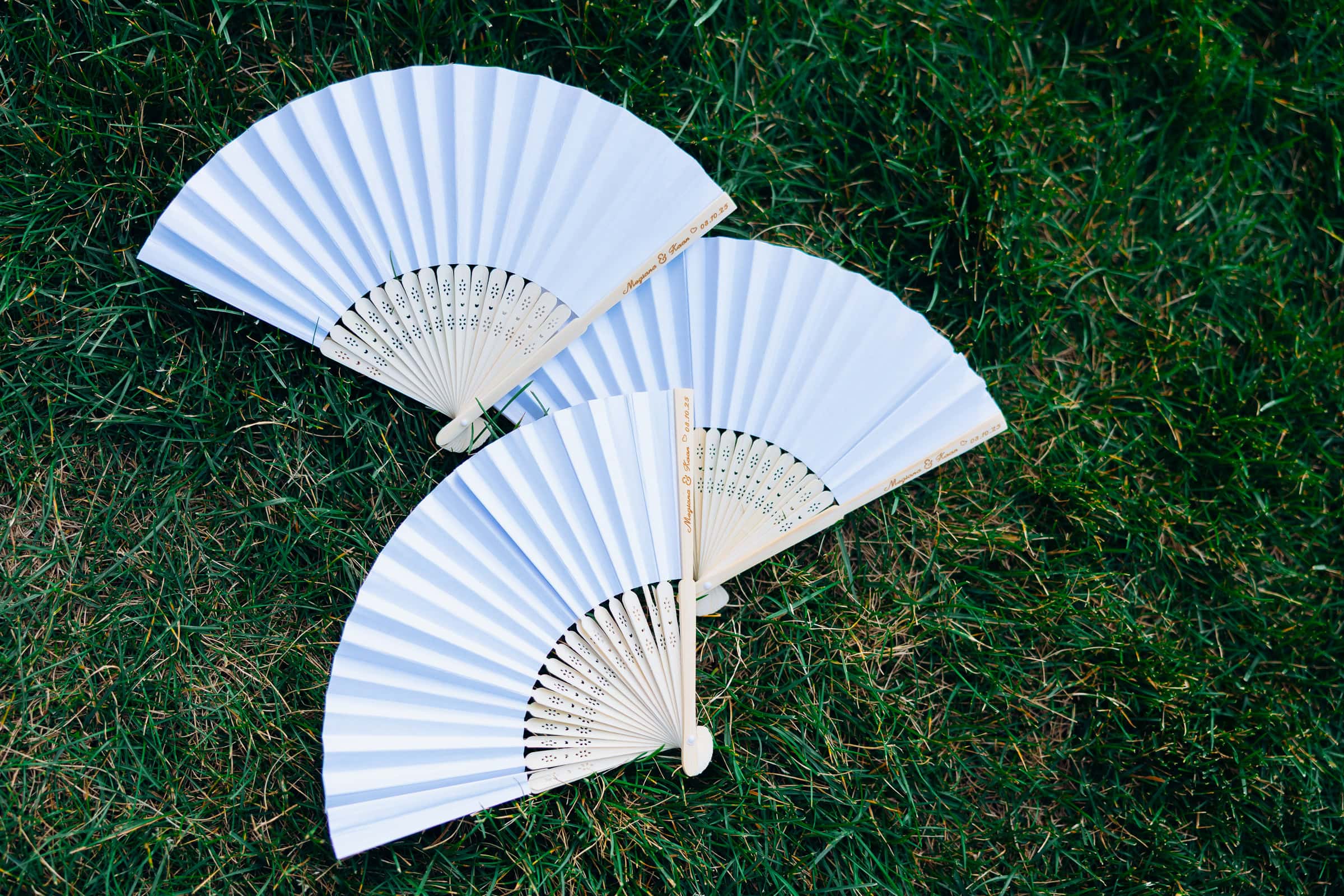 Two white folding fans with wooden ribs engraved with couple's names and wedding date, placed on green grass.