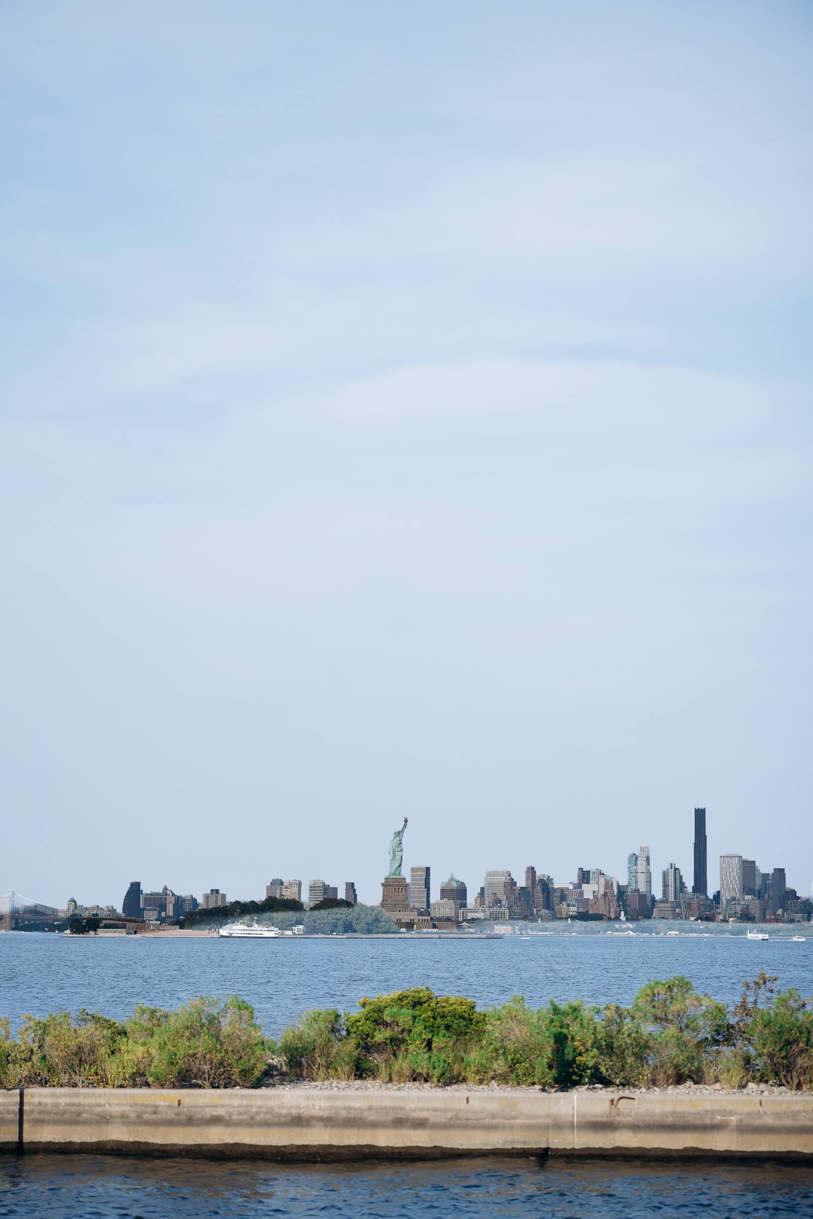 View of Manhattan skyline with Statue of Liberty in center, seen across water with greenery and flagpoles in foreground, Hudson House Jersey City
