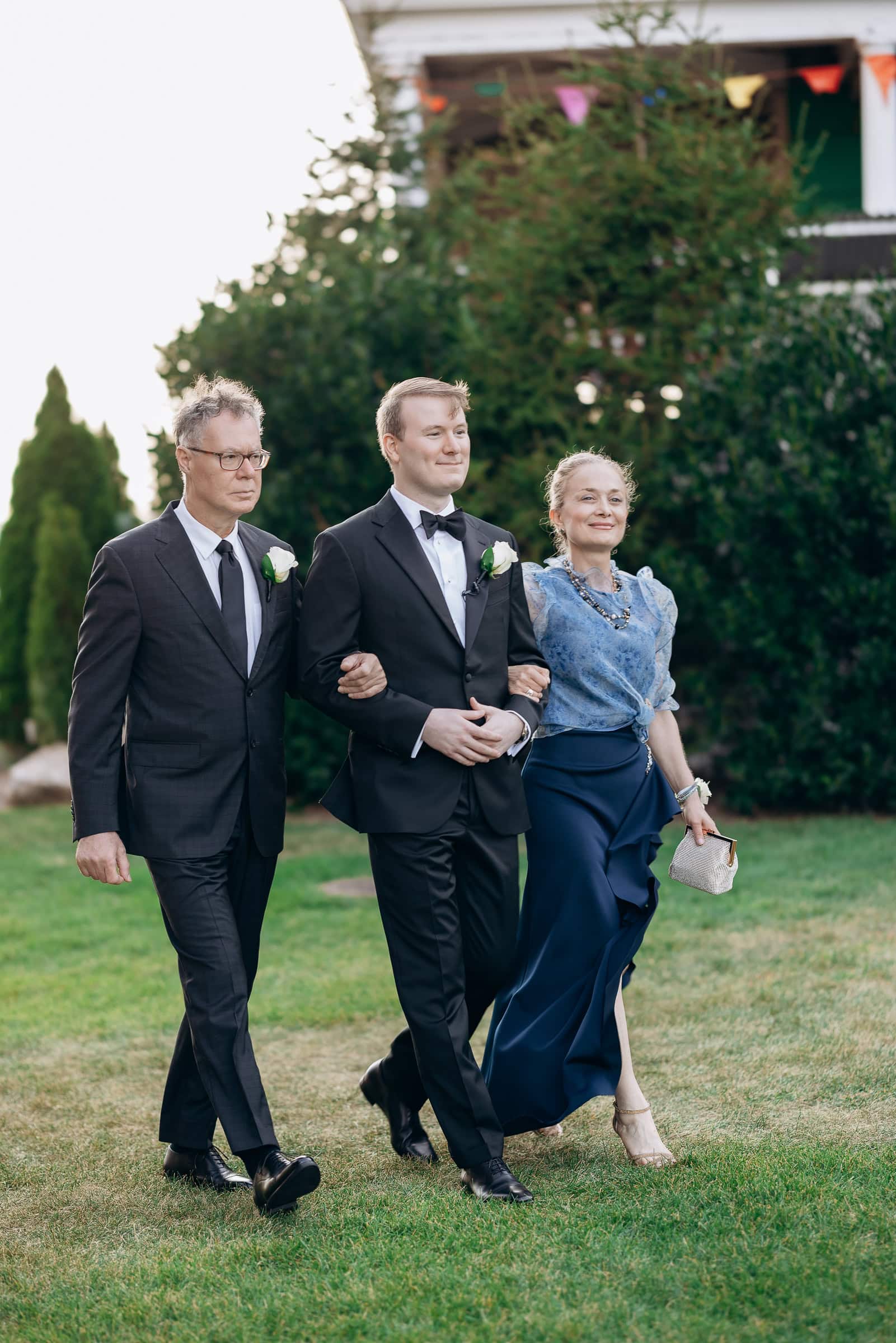 Groom in black tuxedo walking arm-in-arm with his mother in a blue gown and father in a dark suit on grass at Hudson House, Jersey City
