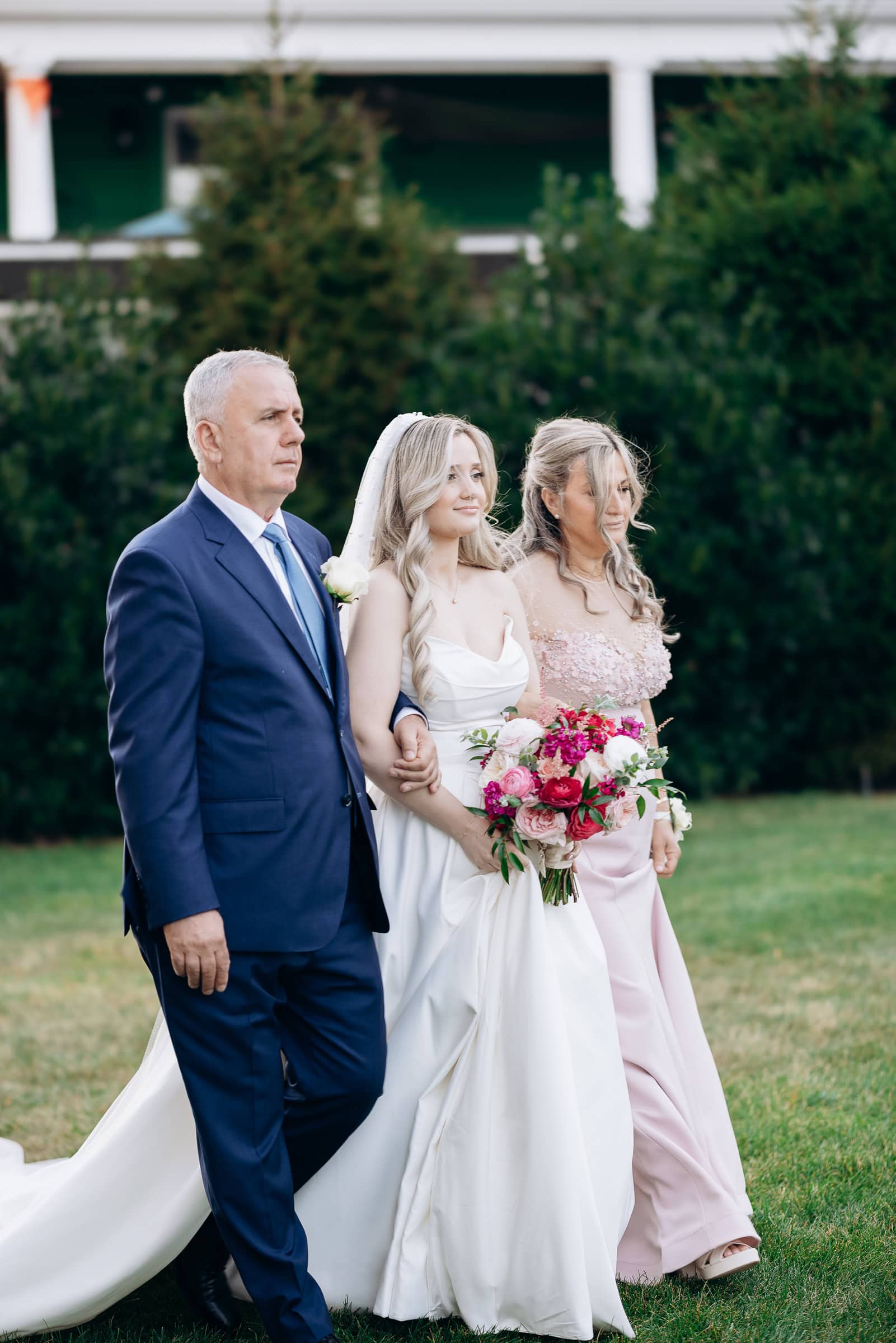 Bride in white gown walking arm-in-arm with her father in a navy suit and mother in a blush pink dress, holding a bouquet of pink and red flowers outdoors at Hudson House, Jersey City.