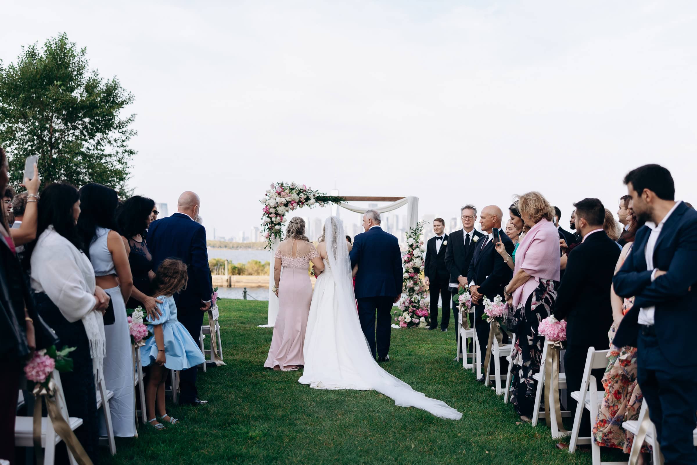 Bride walking down grassy aisle with escort, guests on both sides, floral arch and waterfront view at Hudson House, Jersey City, NJ.