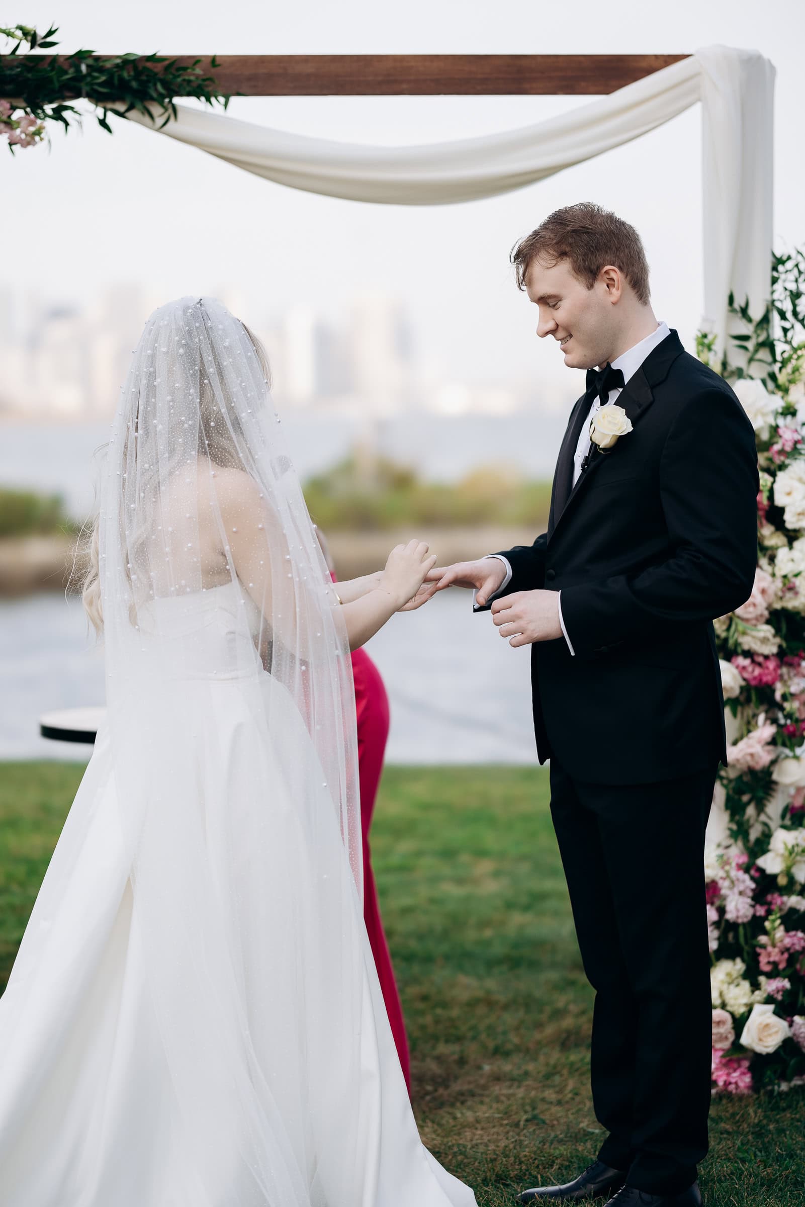 Groom placing wedding ring on bride's finger under white draped arbor with floral arrangements at Hudson House, Jersey City, NJ.