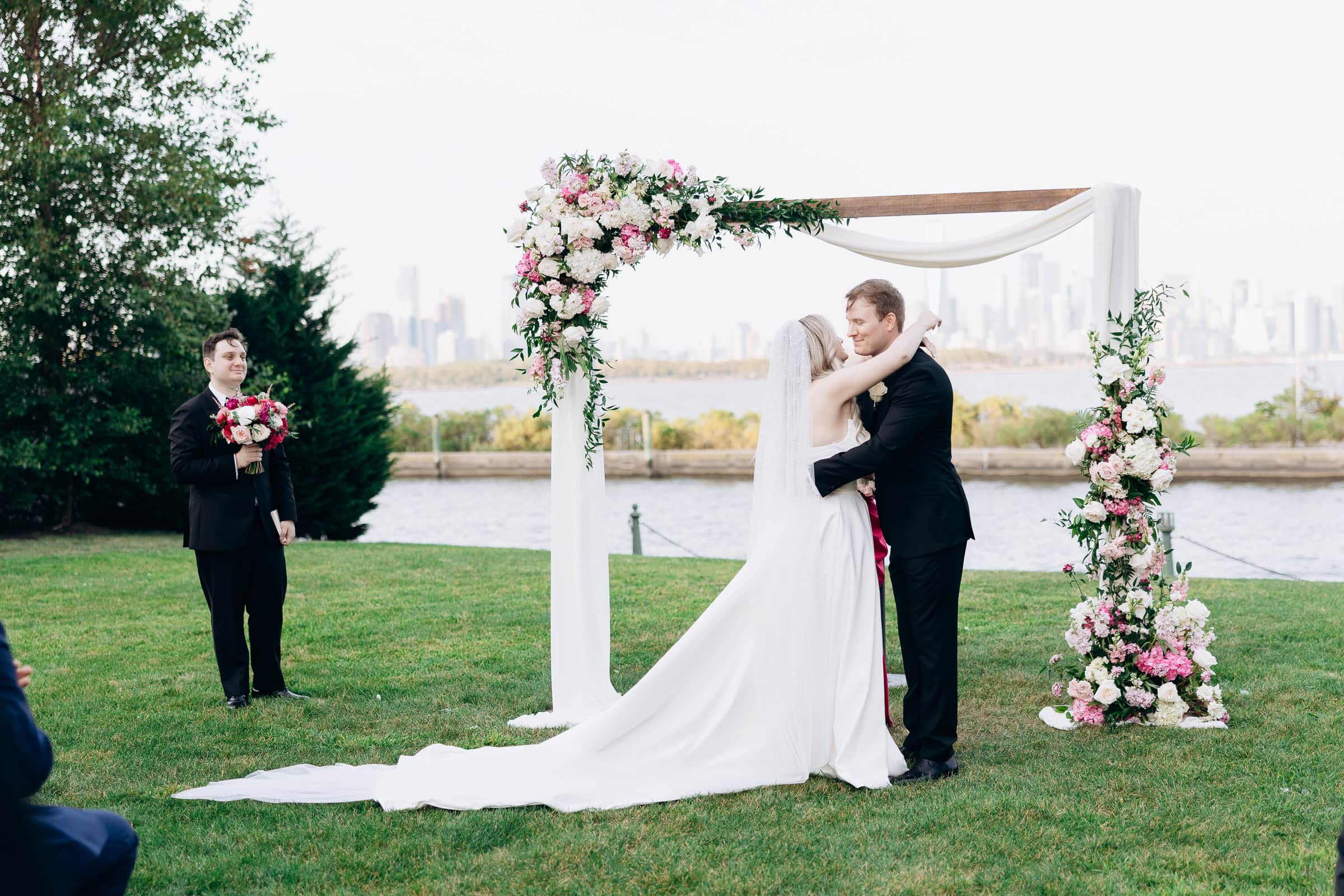 Bride and groom embracing under a floral arch by the water with Manhattan skyline in the background at Hudson House, Jersey City, NJ.