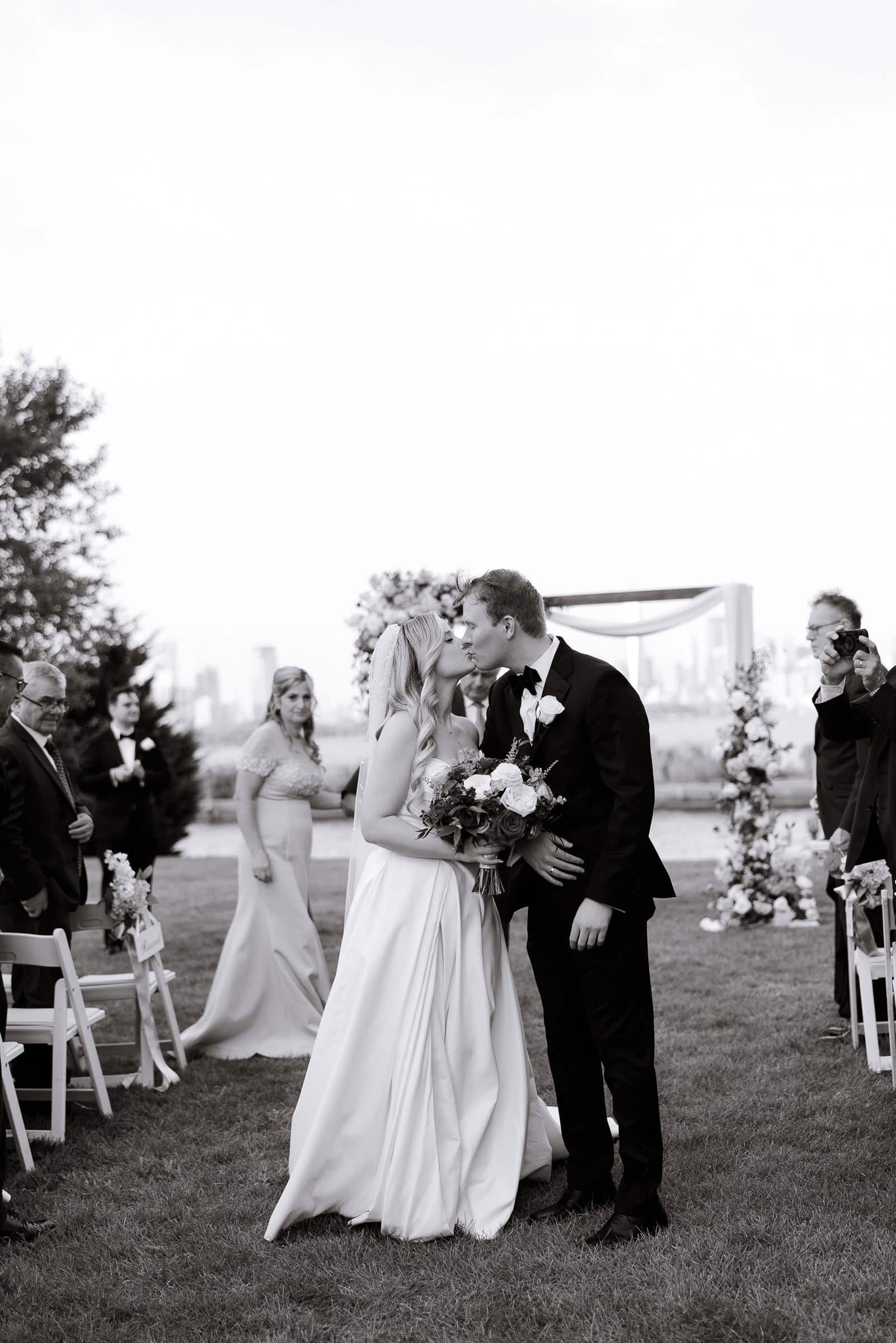 Bride and groom kissing outdoors at Hudson House wedding ceremony with floral arch and guests in background