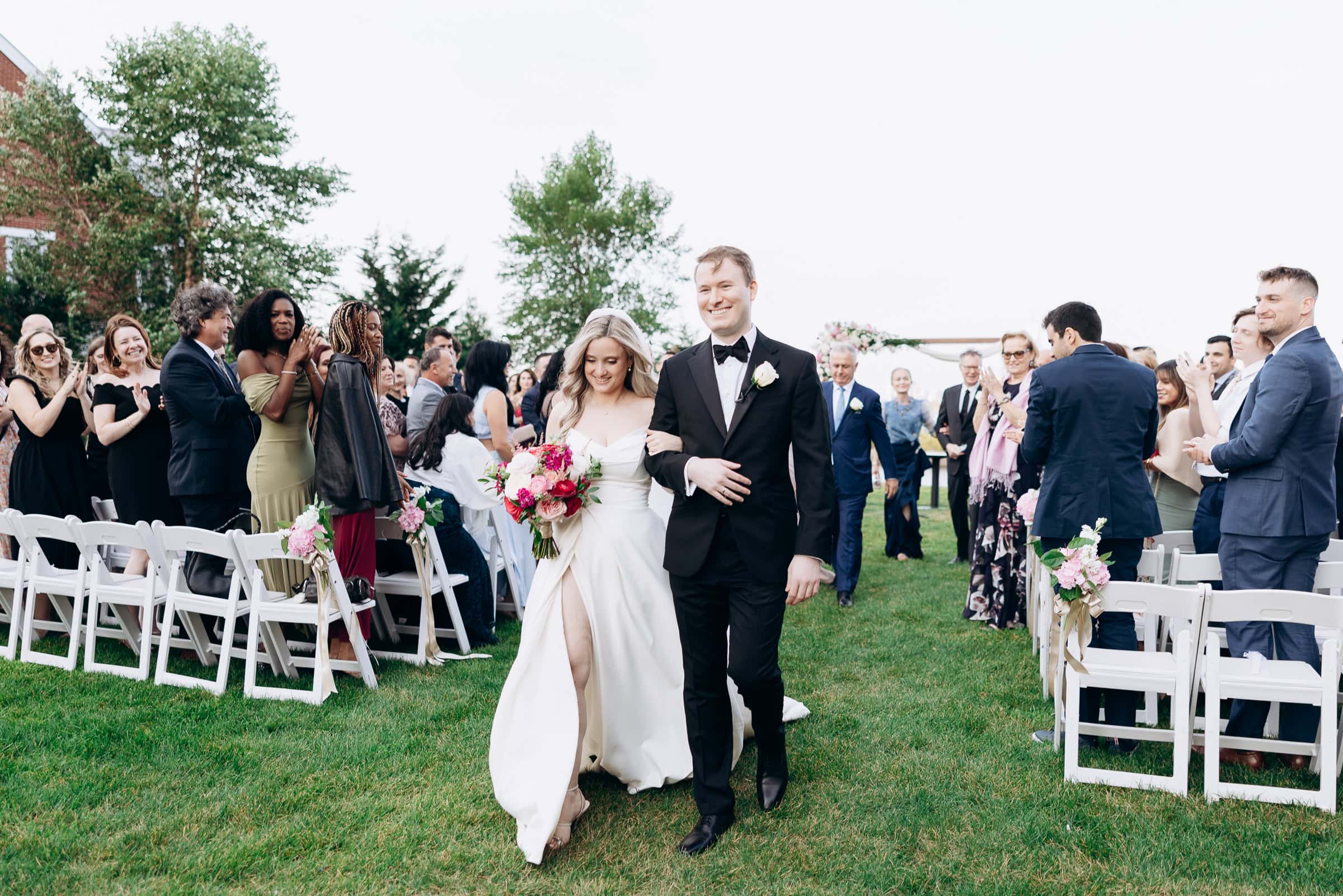 Bride in satin gown holding pink and white bouquet, walking arm-in-arm with groom in black tuxedo down grassy aisle, guests applauding on both sides at Hudson House, Jersey City, NJ.