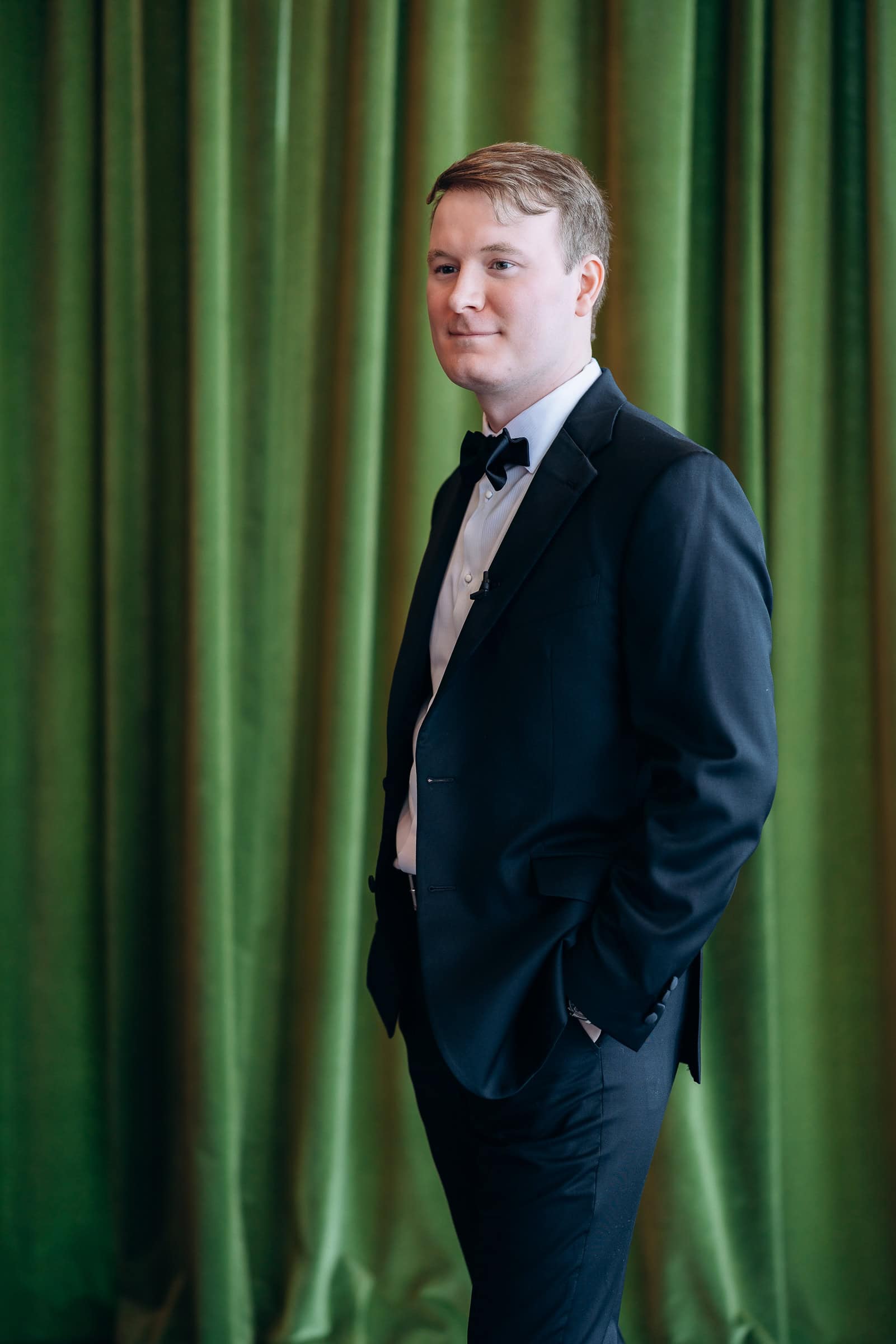 Man in black tuxedo with bow tie standing in front of green velvet curtains, Hudson House, Jersey City, NJ.