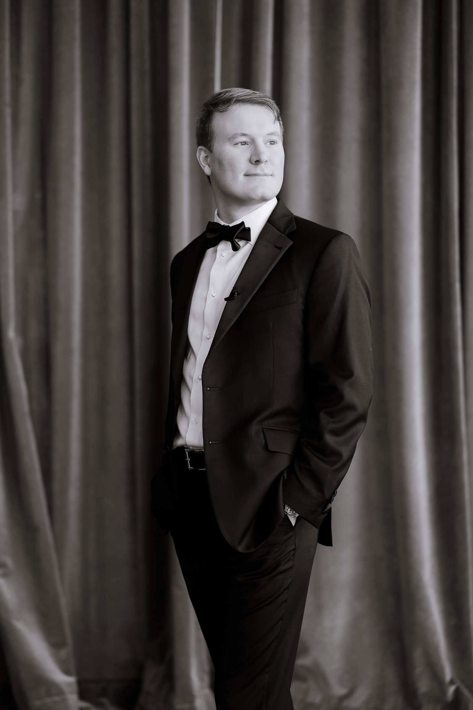 Portrait of a groom in a black tuxedo and bow tie standing in front of green velvet curtains with hands in pockets.