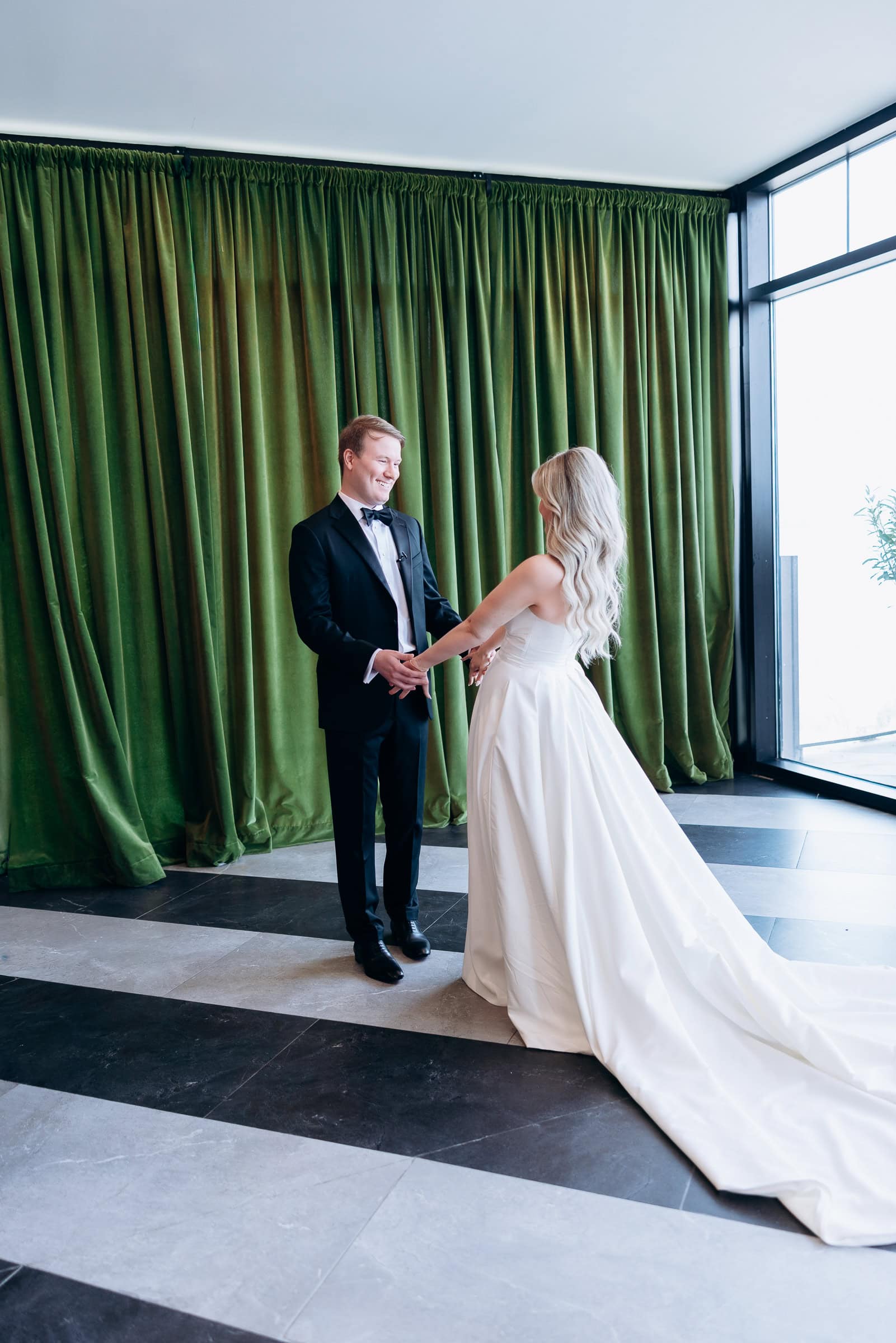 Groom in black tuxedo holding hands with bride in ivory wedding dress in front of green velvet curtains near large window at Hudson House, Jersey City.