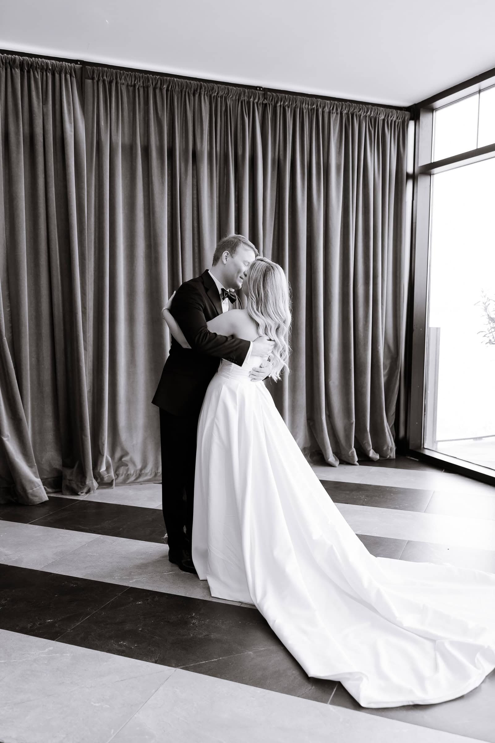 Bride and groom embracing indoors by large windows with olive-green velvet curtains at Hudson House, Jersey City, NJ.