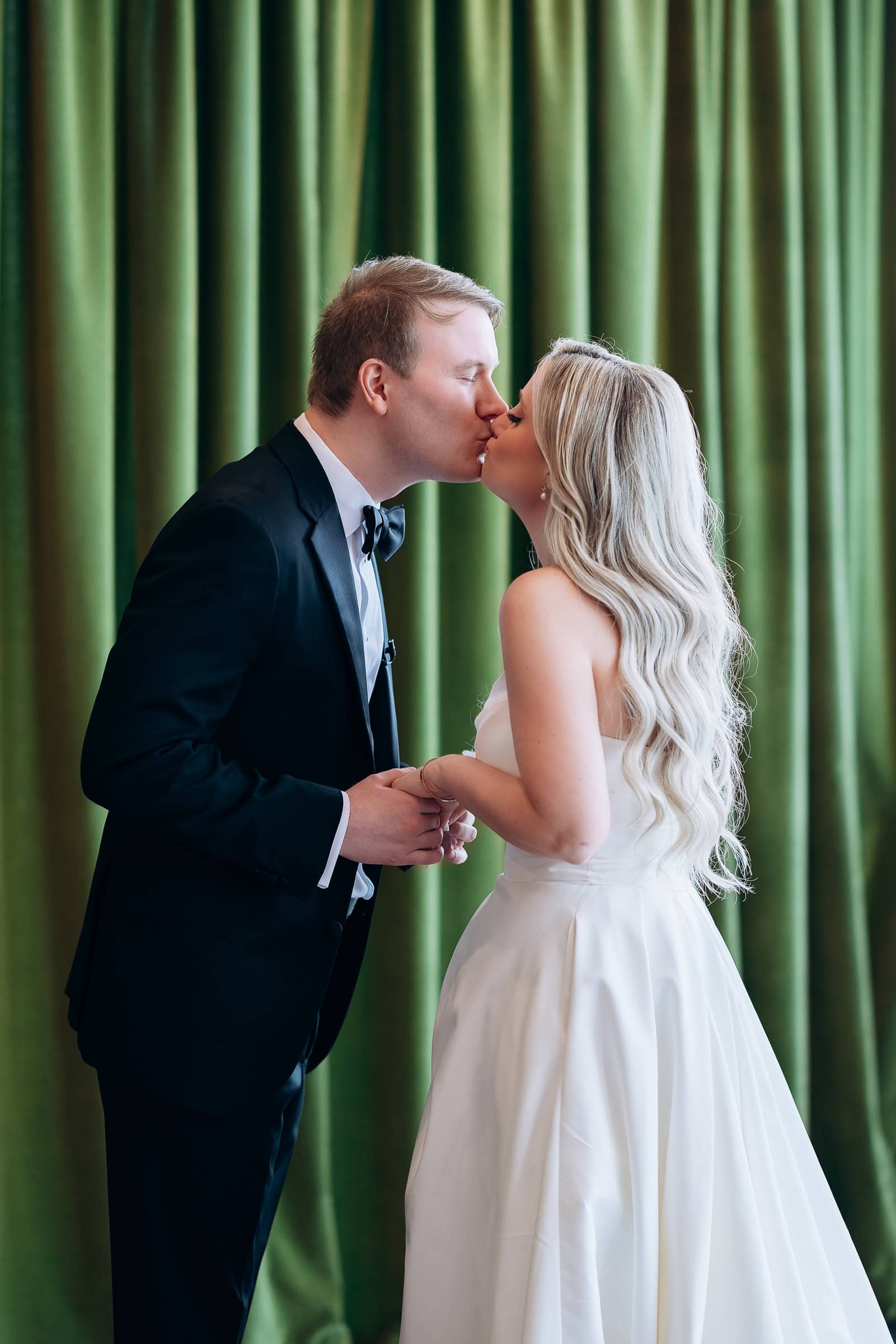 Bride and groom kissing indoors, groom in black tuxedo, bride in white gown with long blonde hair, olive green curtains in background, Hudson House Jersey City