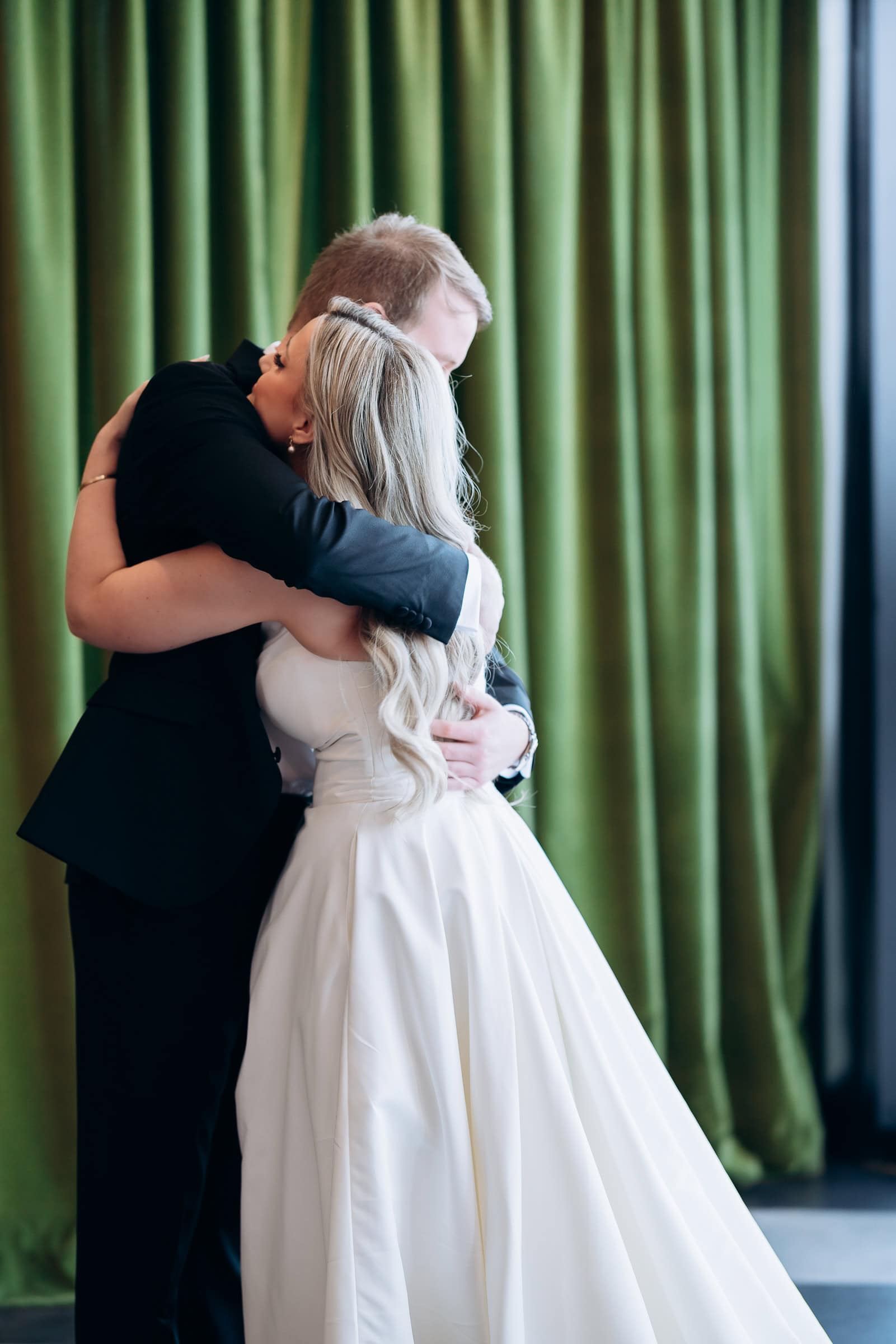 Groom in black suit embracing bride in white gown with long blonde hair, set against olive green curtains at Hudson House, Jersey City.