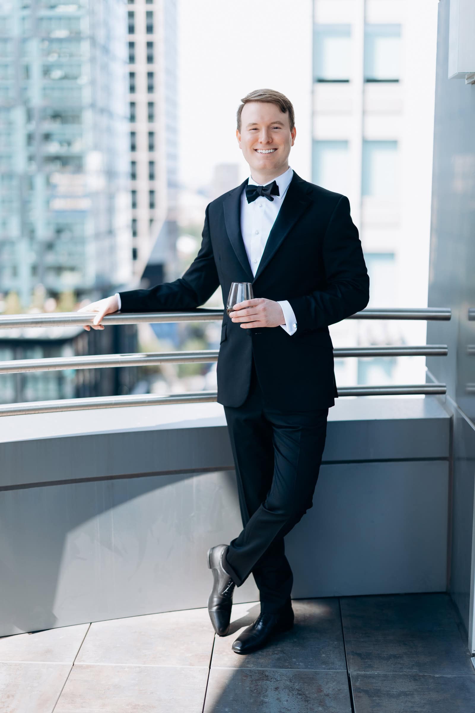 Man in black tuxedo and bow tie holding a glass of whiskey, leaning on balcony railing with city buildings blurred in background.