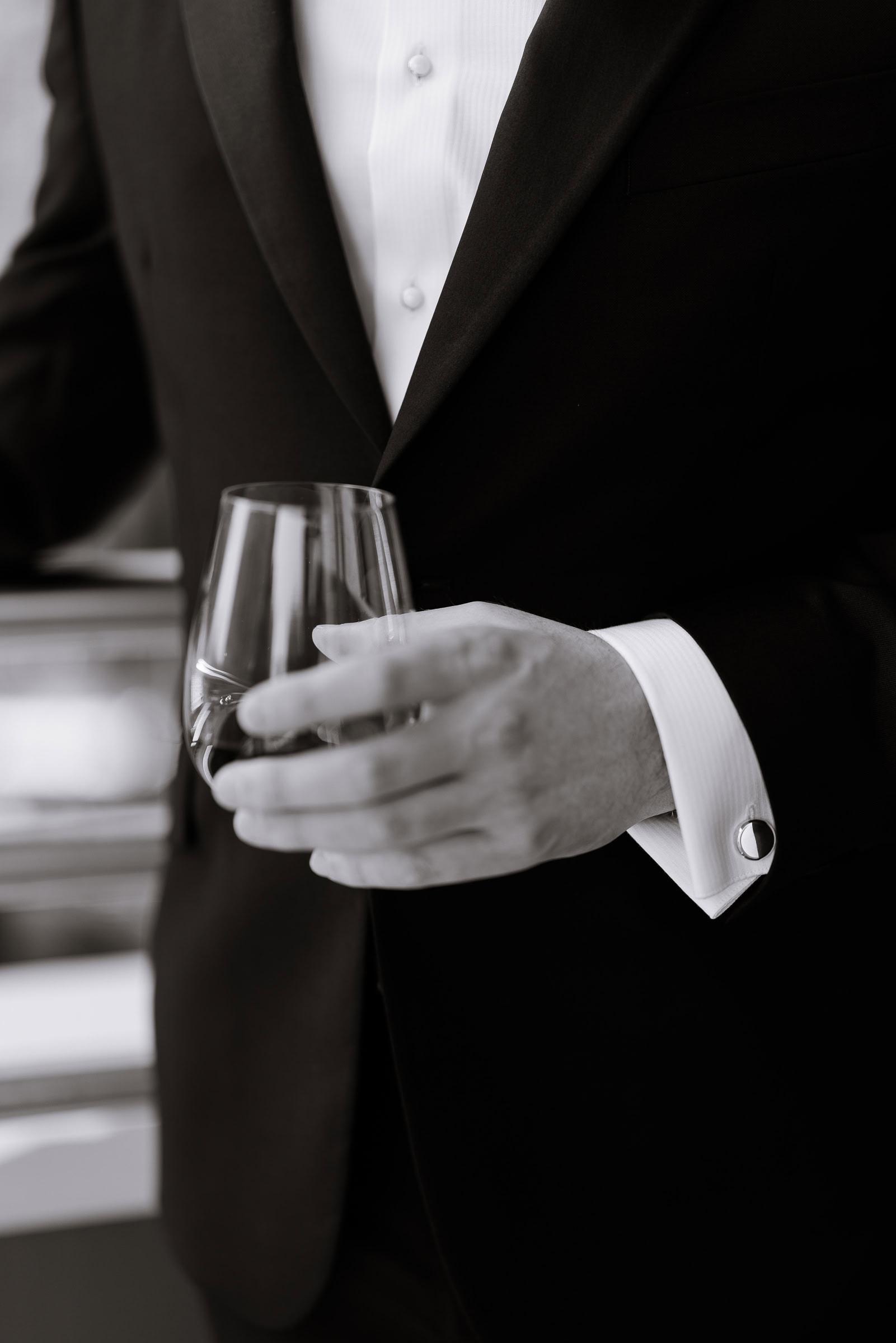Close-up of a groom wearing a black tuxedo and white shirt with pearl buttons and crystal cufflinks, holding a whiskey glass.