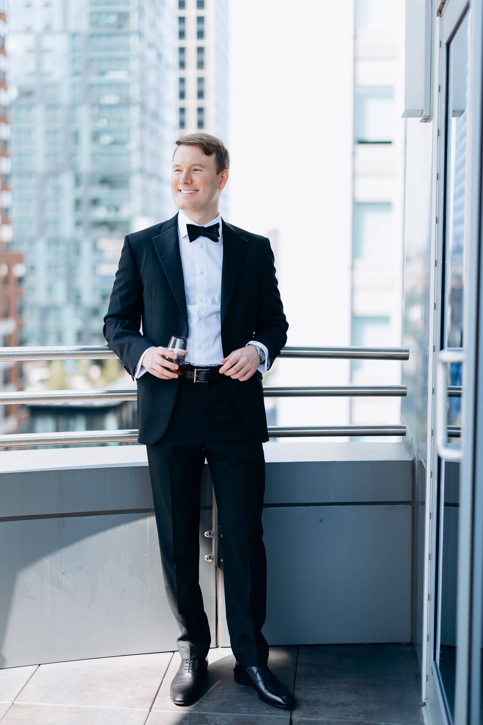 Man in black tuxedo with bow tie holding a glass of whiskey on a balcony overlooking city skyscrapers