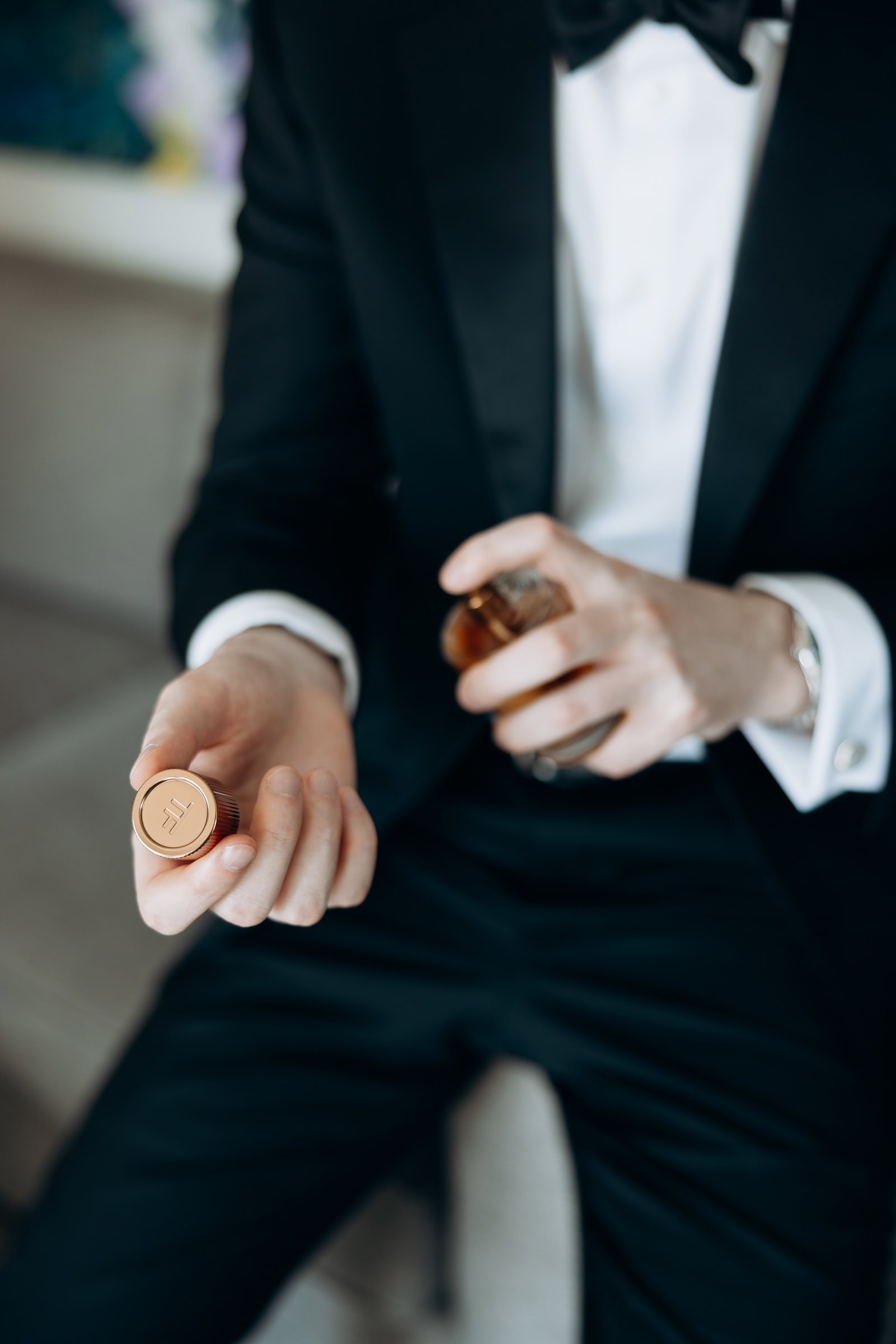 Close-up of a groom in a black tuxedo holding the golden cap of a Tom Ford fragrance bottle, preparing to apply cologne.