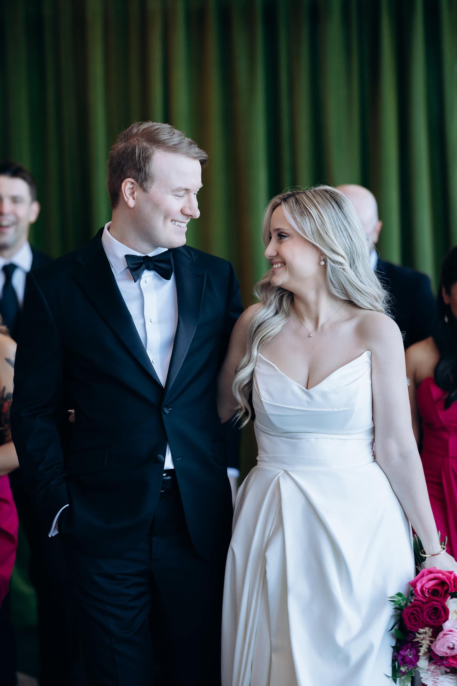 Bride in satin wedding dress holding bouquet of pink and red roses smiling at groom in black tuxedo with bow tie, green curtains in background at Hudson House, Jersey City