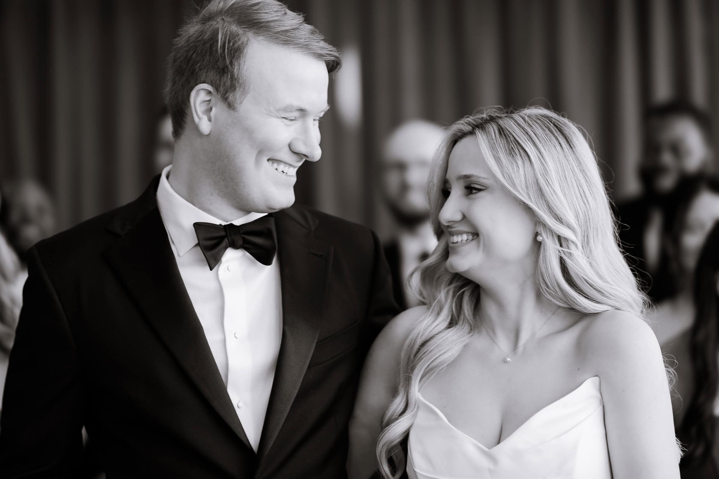 Close-up of a groom in black tuxedo and bride in white gown smiling at each other during wedding at Hudson House, Jersey City, NJ.