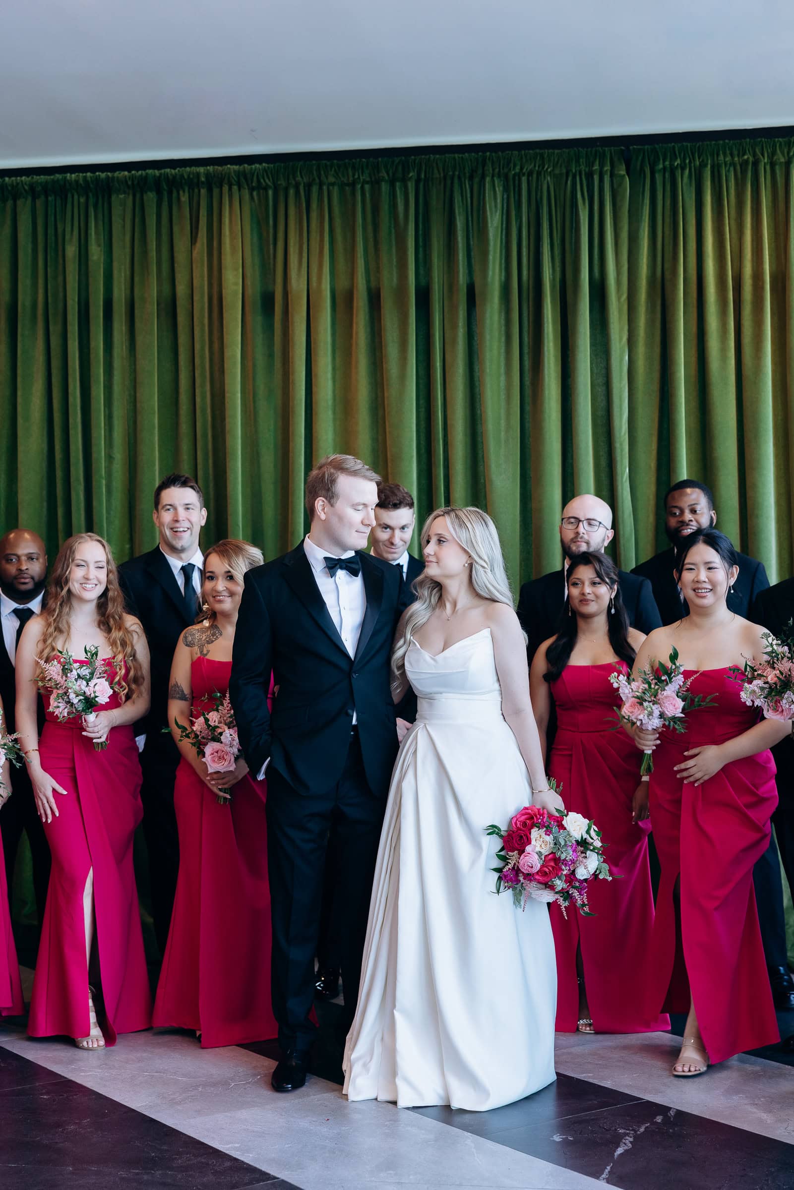 Bride in ivory satin gown and groom in black tuxedo stand closely, surrounded by bridesmaids in burgundy dresses and groomsmen in black tuxedos, with olive green curtains behind.