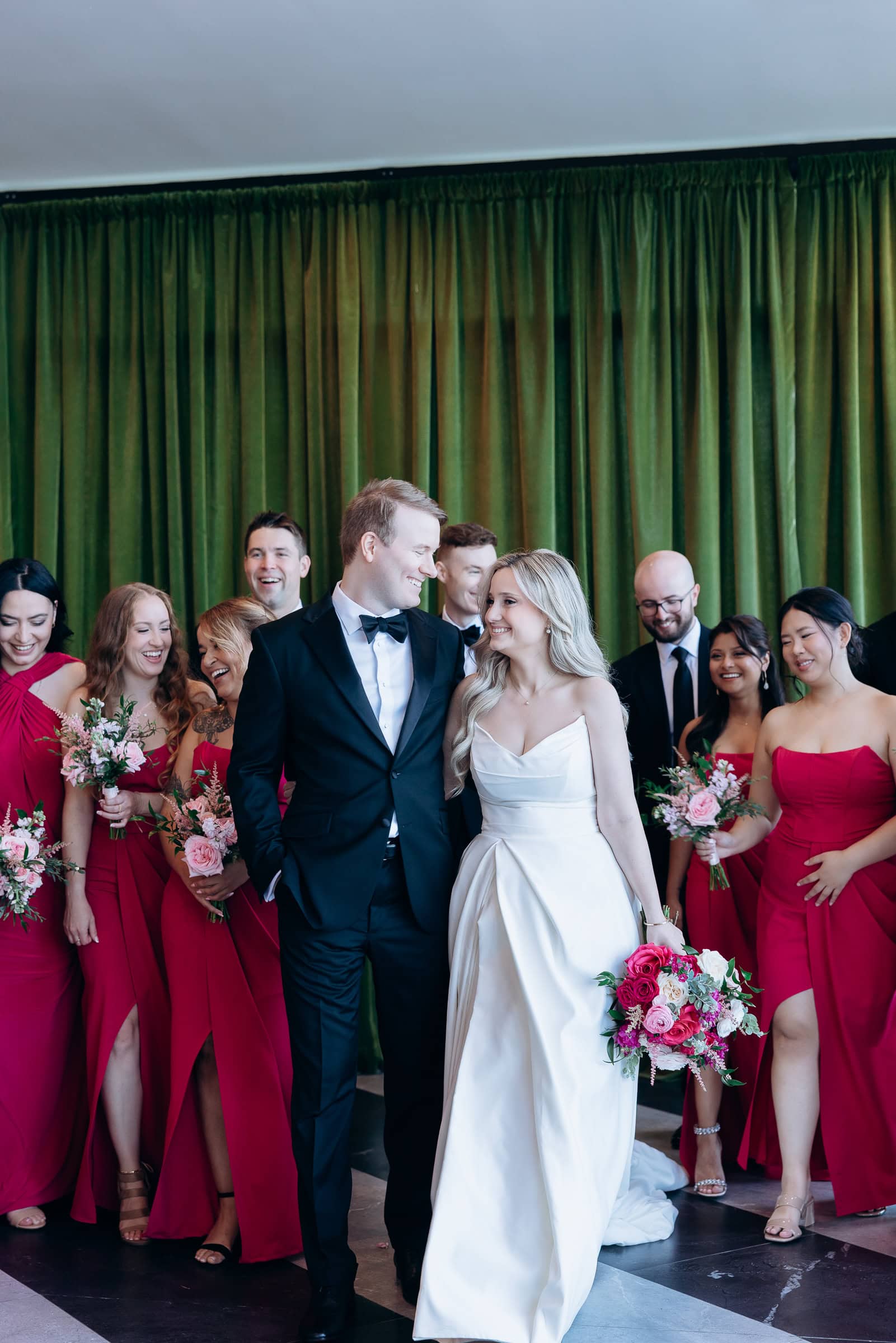 Bride in white gown and groom in black tuxedo smiling at each other, surrounded by bridesmaids in red dresses and groomsmen, indoors with olive green curtains.