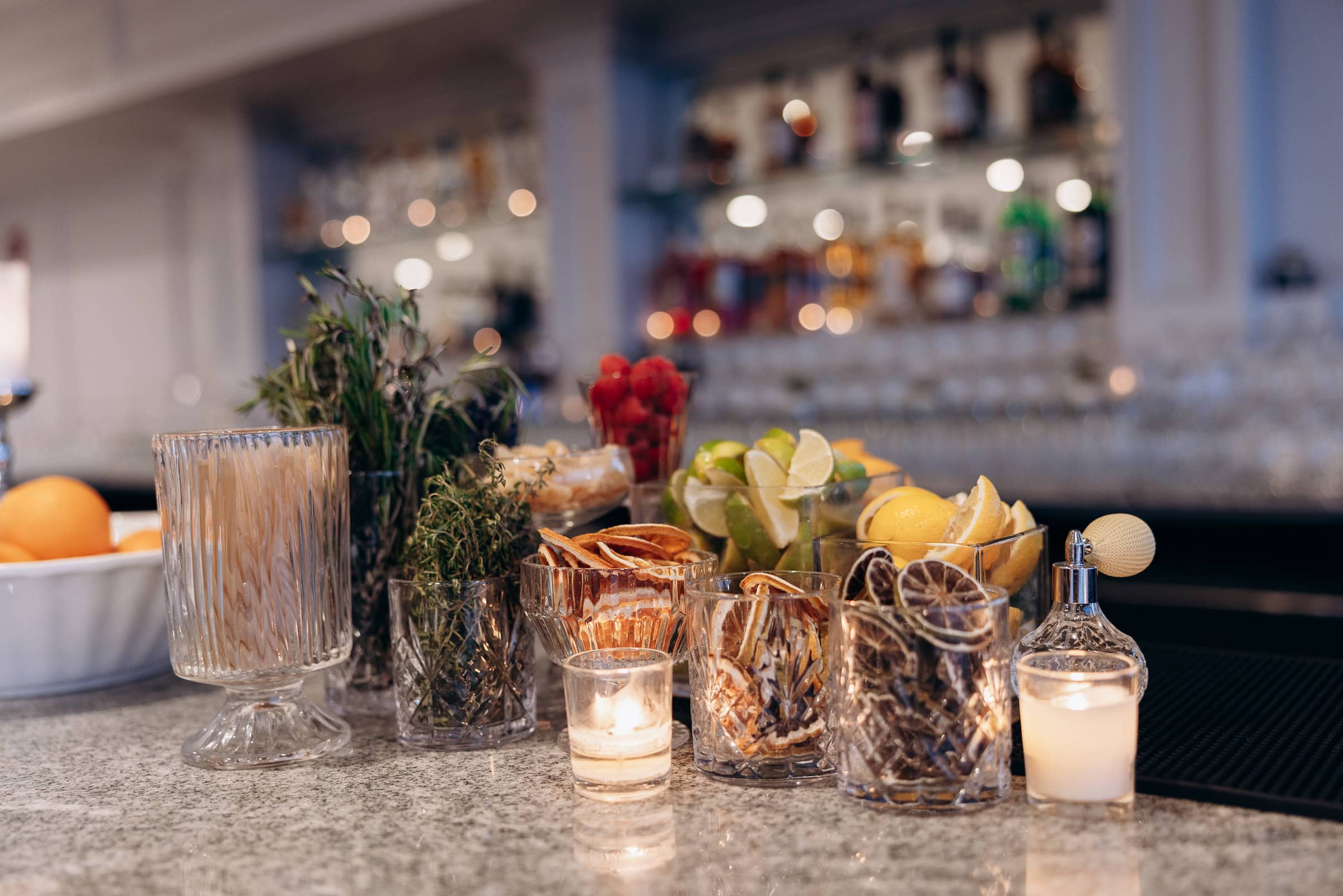 Close-up of cocktail garnishes including dried citrus slices, fresh herbs, lime and lemon wedges, and candles on a bar counter with blurred bottles in the background at Hudson House, Jersey City.